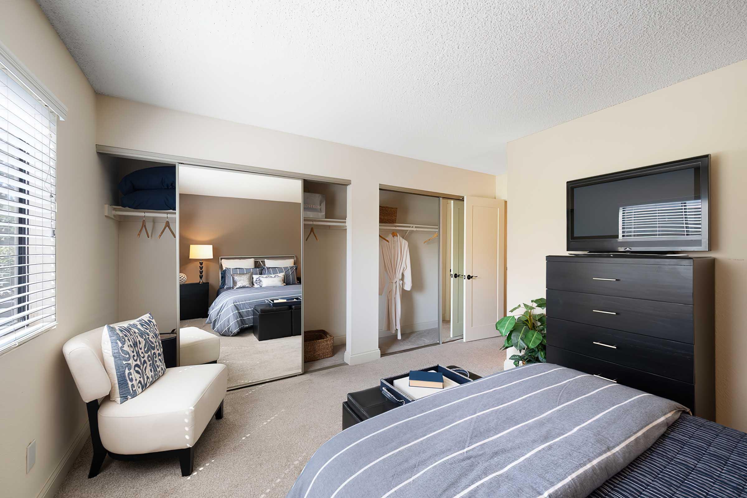 A cozy bedroom featuring a bed with blue and gray bedding, a modern dresser, and a chair. There is a large mirror closet with sliding doors and an open space with a robe hanging. Natural light enters through a window with blinds, and a potted plant adds a touch of greenery to the room.