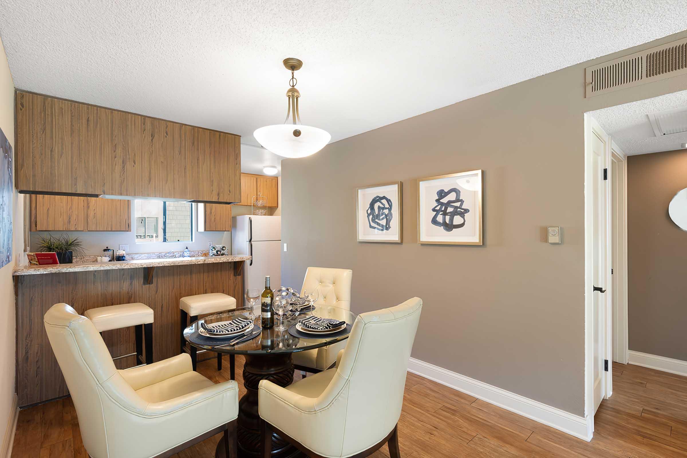 A modern dining area featuring a round wooden table with four cream-colored chairs. The walls are painted a soft gray, adorned with two abstract framed artworks. A breakfast bar with three stools is visible in the background, along with a kitchen area. Natural light illuminates the space.