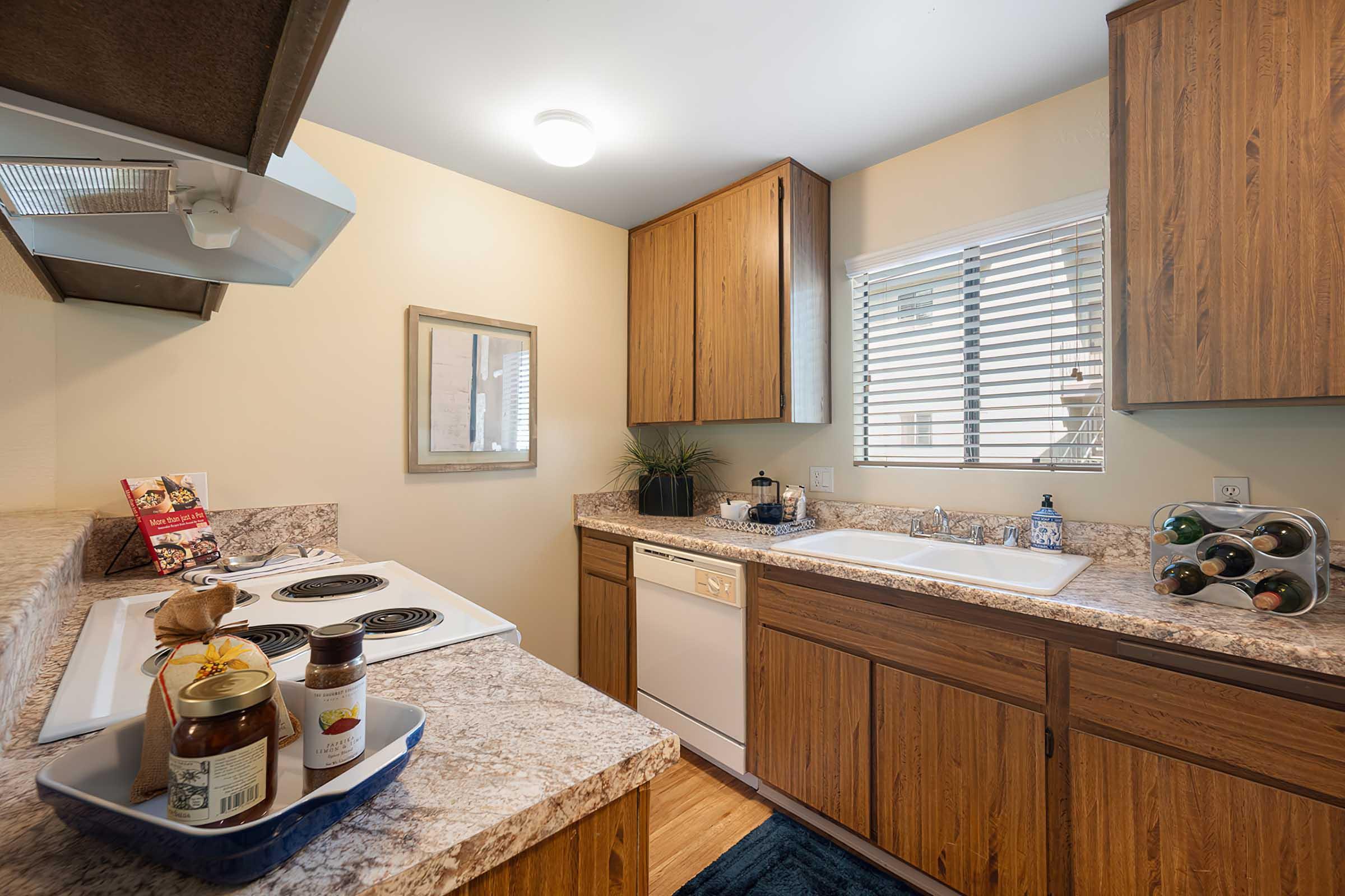A modern kitchen featuring wooden cabinetry, a granite countertop, and stainless-steel appliances. It includes a stove, sink, and dishwasher, with a window allowing natural light. A tray with jars and a wine holder is placed on the counter, adding a cozy touch to the space.