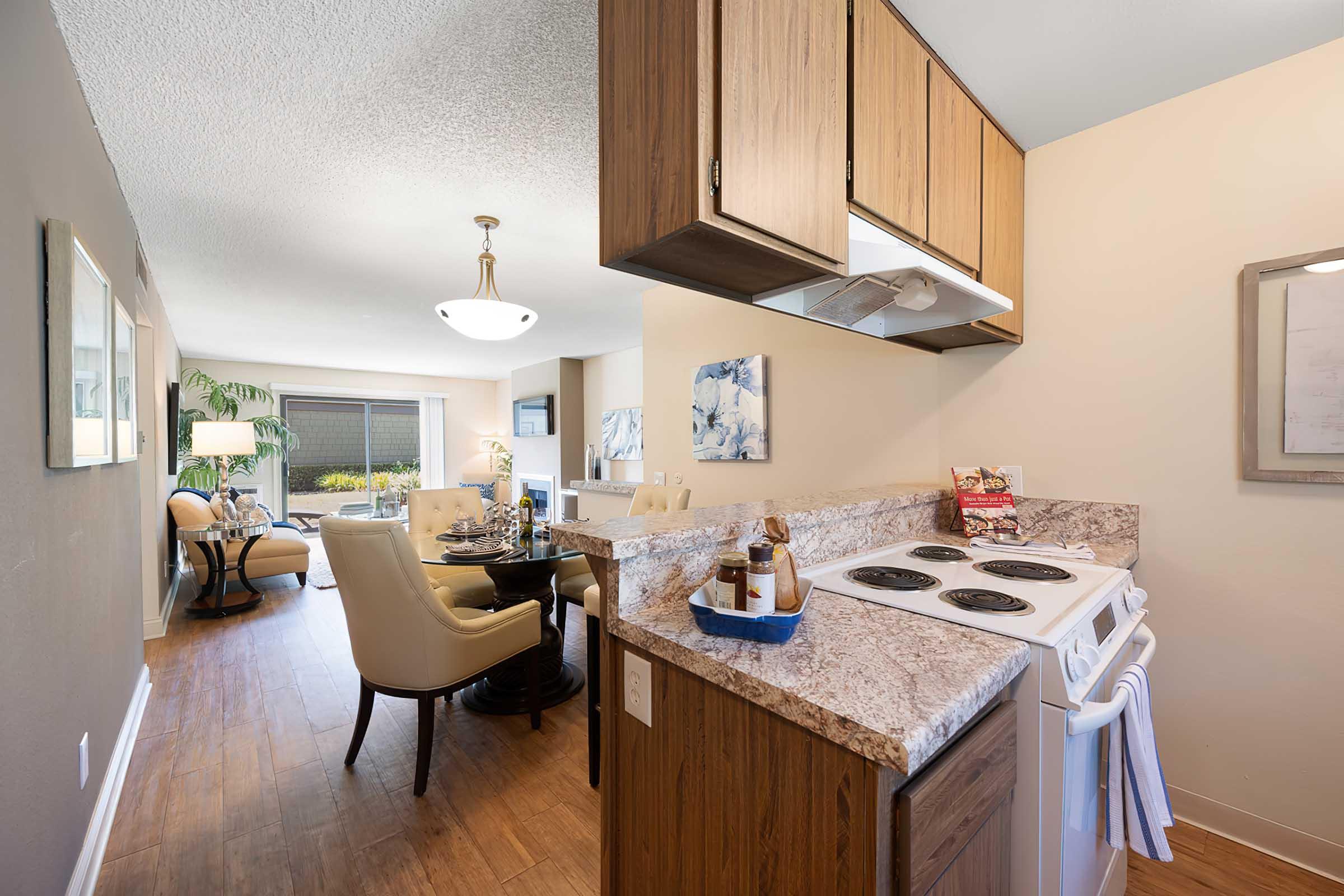 Modern kitchen with a stove and countertop, featuring wood cabinets and a decorative bowl. In the background, a cozy dining area with a round table and chairs is visible, alongside a living space with natural light. Neutral wall colors and stylish decor create a welcoming atmosphere.