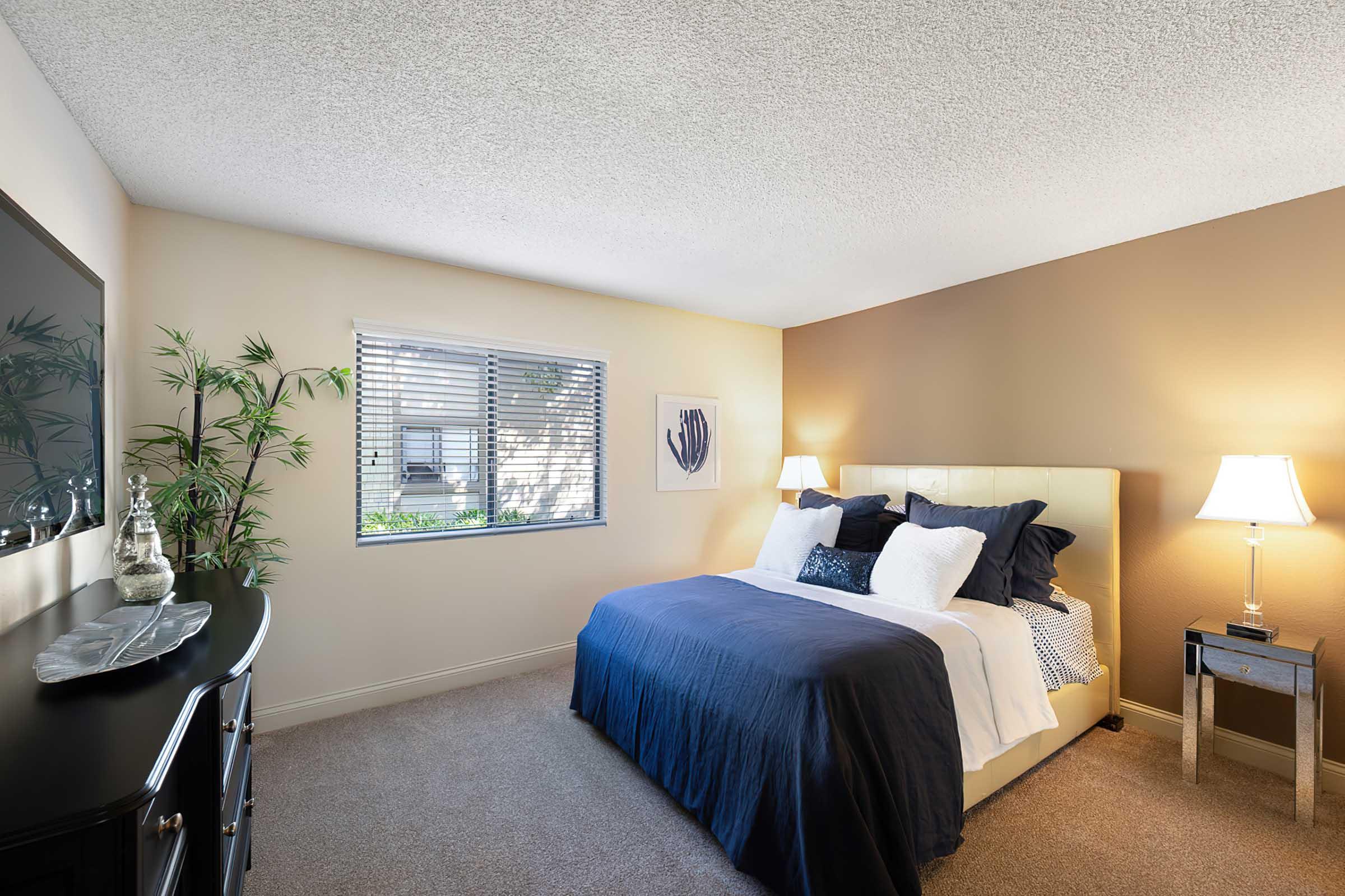 A cozy bedroom featuring a large bed with dark blue and white bedding, two bedside lamps, and a window with blinds that lets in natural light. A decorative plant and a piece of artwork are present, along with a stylish dresser or chest of drawers. The walls are painted in warm tones.