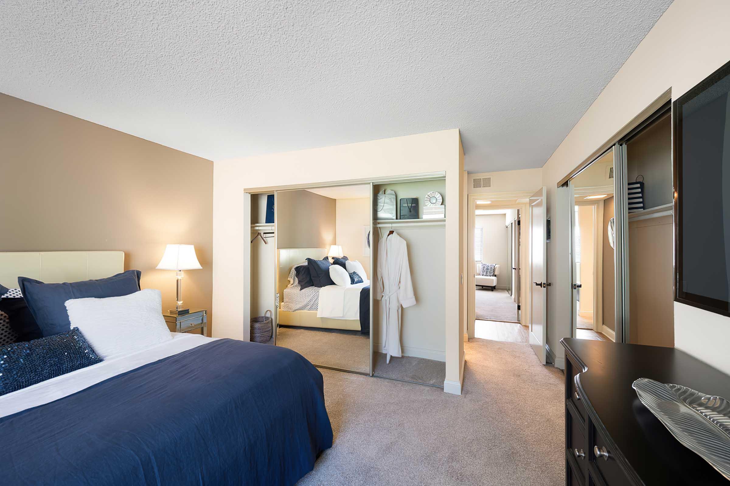 A cozy bedroom featuring a large bed with navy bedding. A bedside lamp provides soft lighting. There are mirrored closet doors reflecting an arrangement that includes a robe and neatly organized storage. The walls are a warm beige, complementing the plush carpet and creating a welcoming atmosphere.