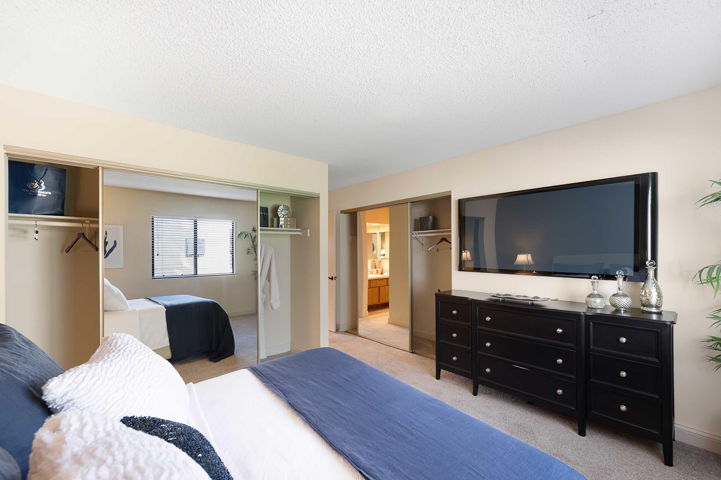 A cozy bedroom featuring a bed with a blue and white comforter, a dark wooden dresser, and a wall-mounted TV. Two open closets are visible, and a window brings in natural light. A small hallway leads to a bathroom, creating a spacious and inviting atmosphere.