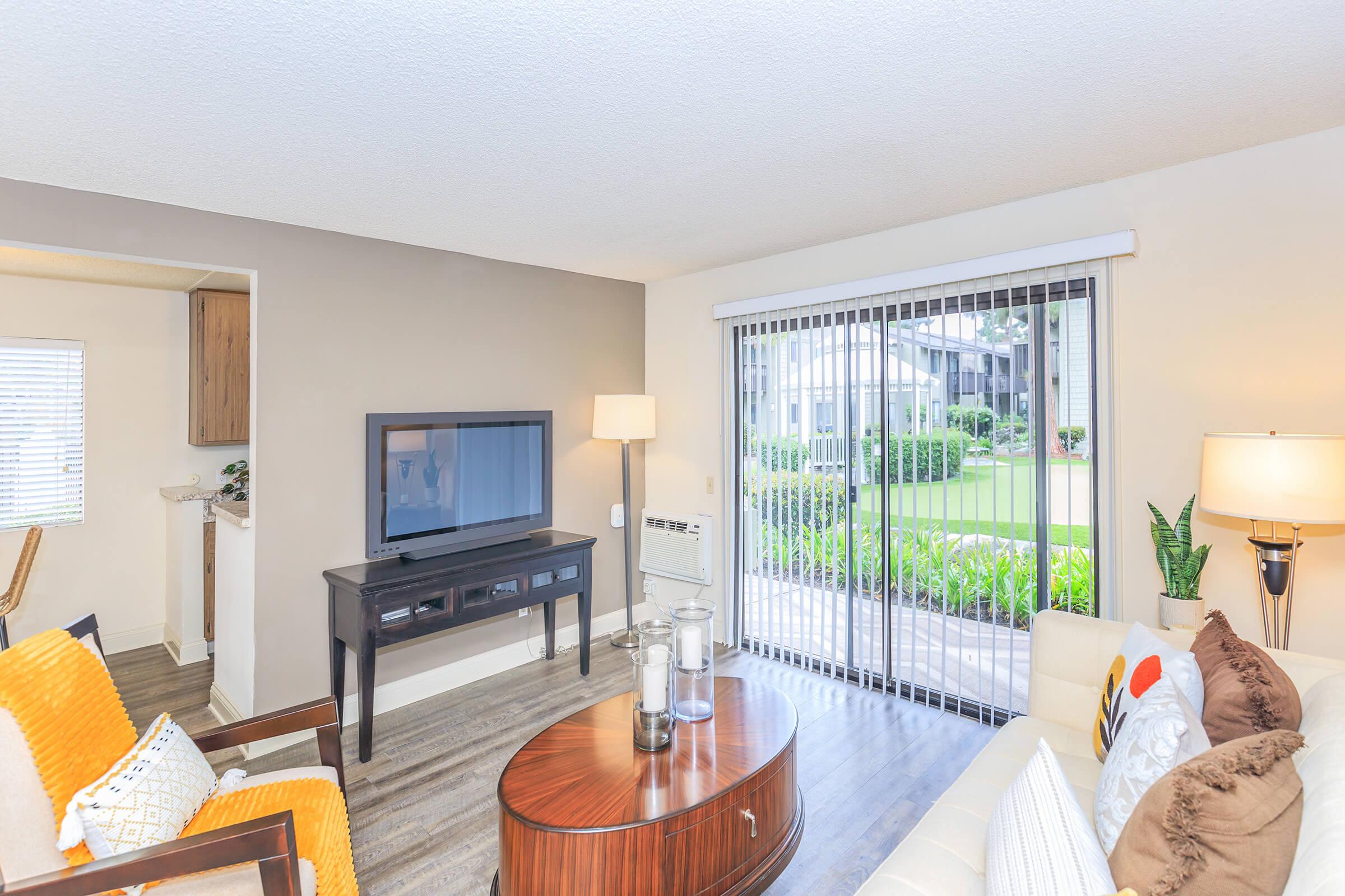 Modern living room with a light-colored sofa, a round wooden coffee table, and a flat-screen TV on a dark console. A lamp provides warm lighting, and sliding glass doors open to a green outdoor area. Bright pillows add color, enhancing the cozy atmosphere.