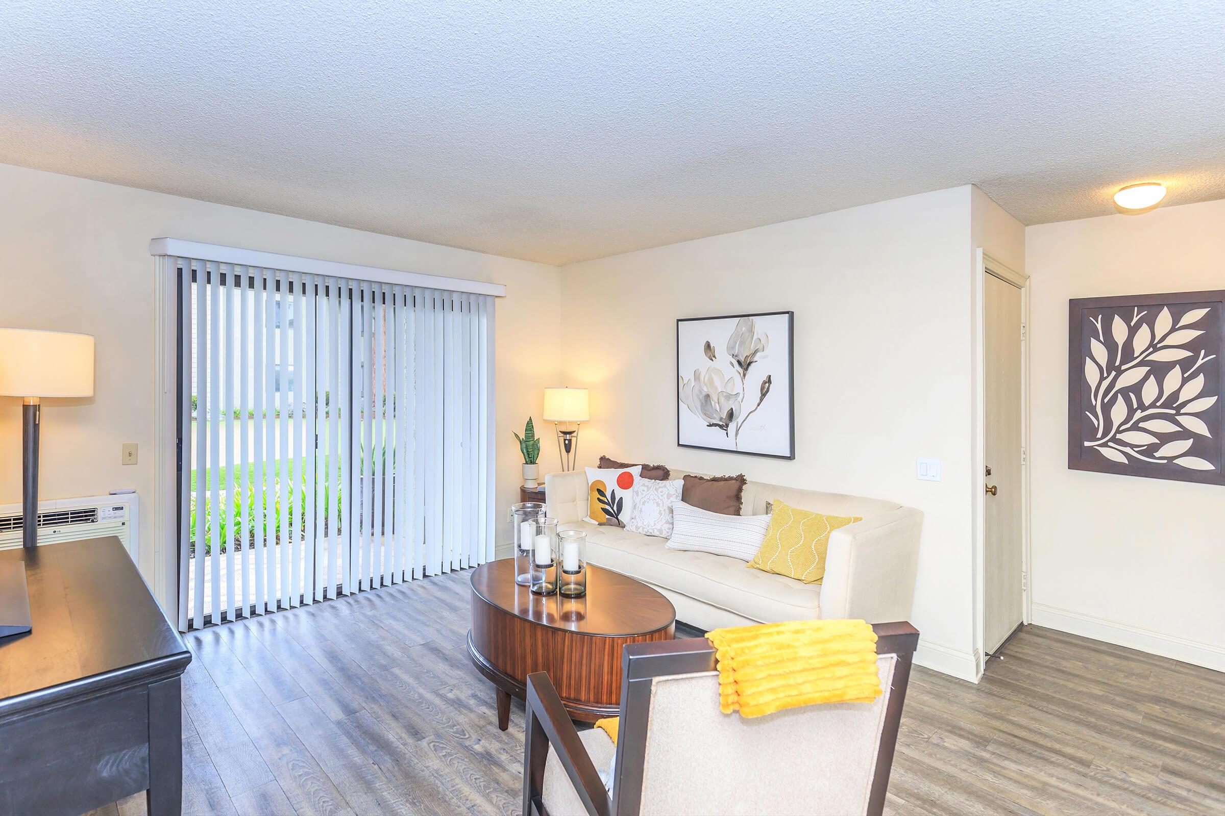 A cozy living room featuring a light-colored couch with decorative pillows, a round coffee table with candles, and a dark wooden desk. Large sliding glass doors with vertical blinds allow natural light, leading to an outdoor view. A potted plant adds a touch of greenery to the space.