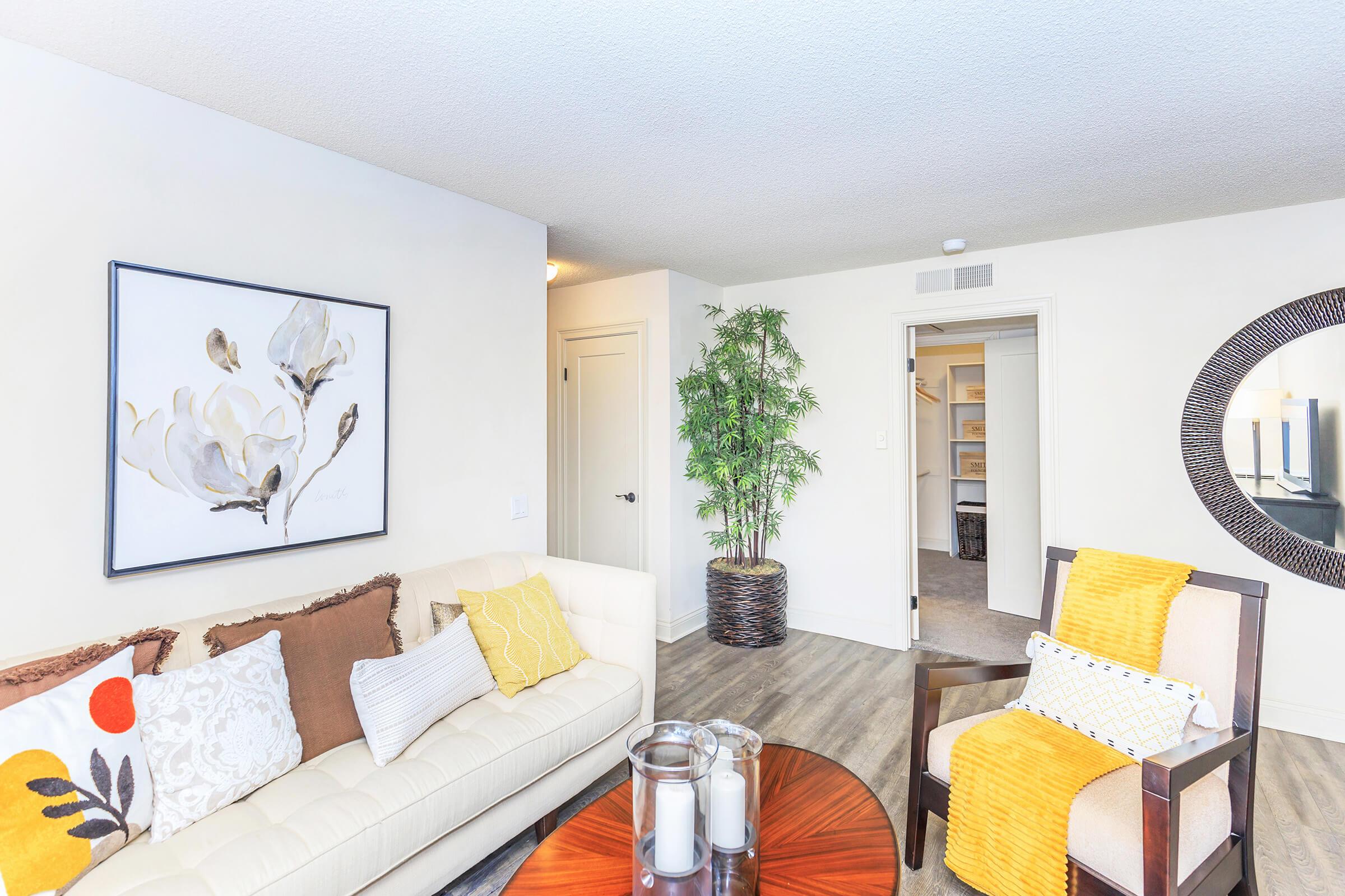 A cozy living area featuring a white sofa with decorative pillows, a wooden coffee table with candles, a chair with a yellow throw, a potted plant, and wall art depicting flowers. The space has light-colored walls and a doorway leading to an adjacent room, creating a bright and inviting atmosphere.
