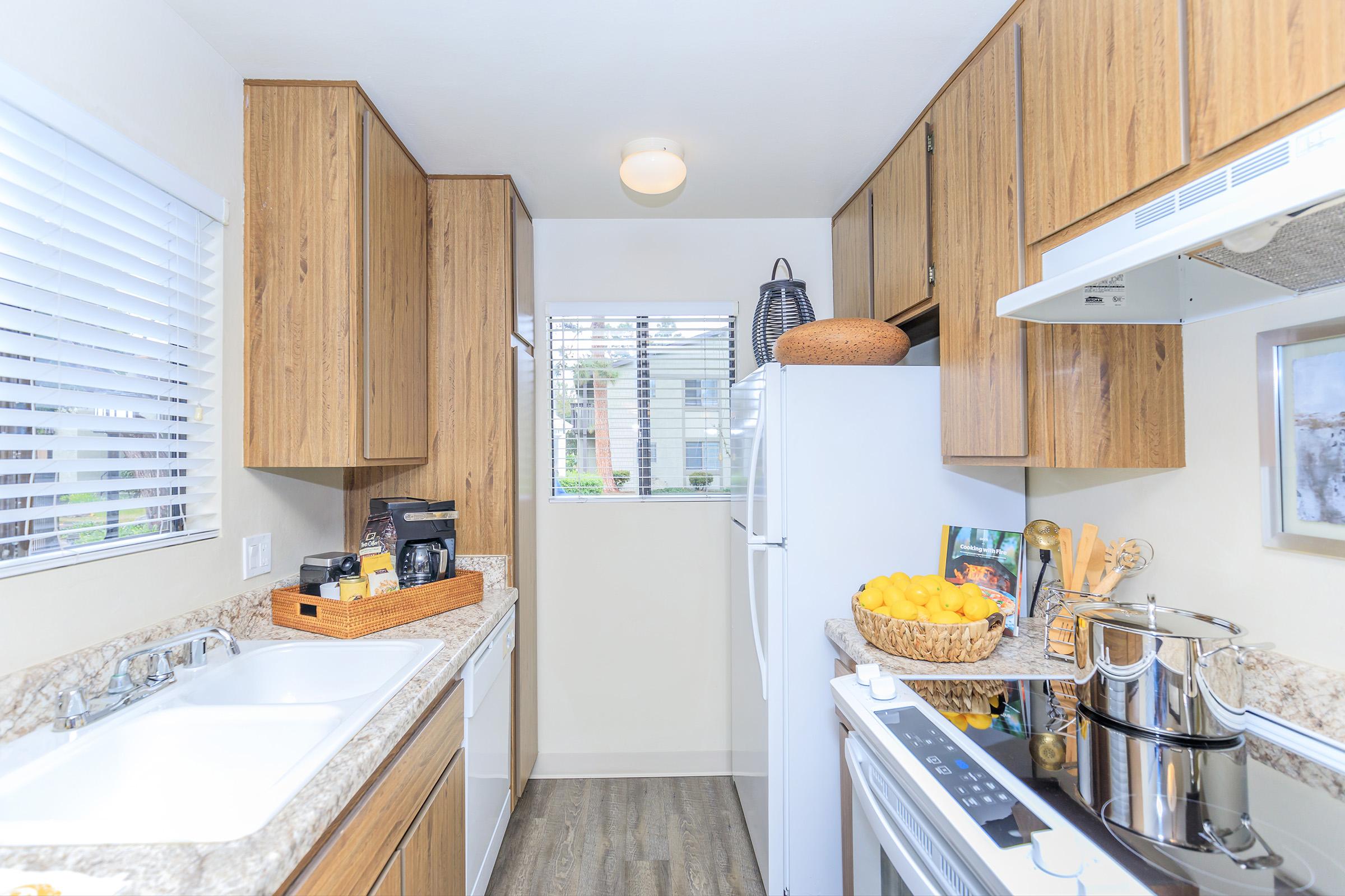 A modern kitchen with wooden cabinets and a granite countertop. Features a double sink, a white refrigerator, and a stove with pots. A basket of lemons sits on the counter, and there is a window providing natural light. The space is tidy and well-organized, showcasing a cozy and functional design.