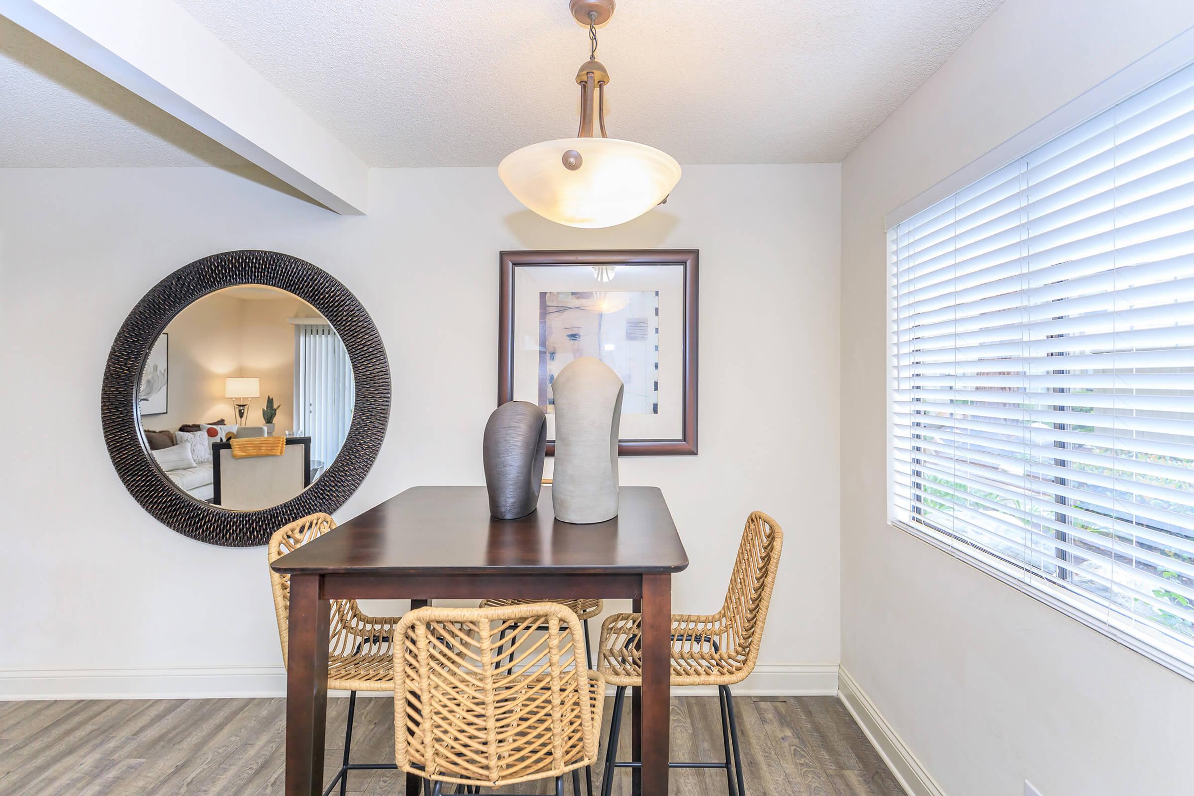 A cozy dining area featuring a wooden table with two decorative vases on it, surrounded by four woven chairs. A circular mirror hangs on the wall, reflecting a softly lit room with a window featuring white blinds. The walls are light-colored, contributing to a warm and inviting atmosphere.