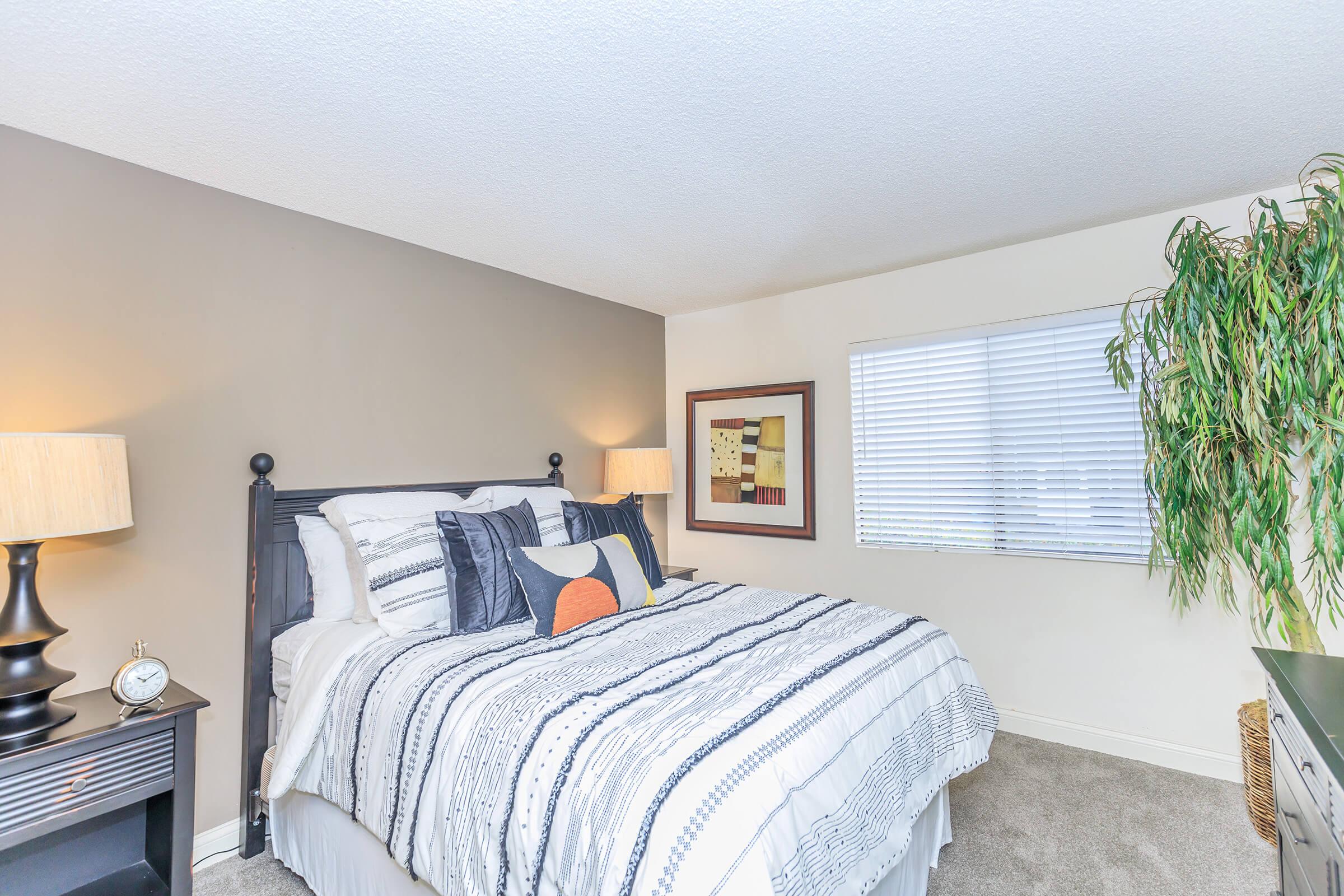 A cozy bedroom featuring a black wooden bed with a striped comforter and decorative pillows. Two bedside lamps illuminate the space. A framed abstract artwork hangs on the wall next to a window with closed blinds, and a tall plant adds a touch of greenery to the room. Soft carpet flooring completes the warm atmosphere.