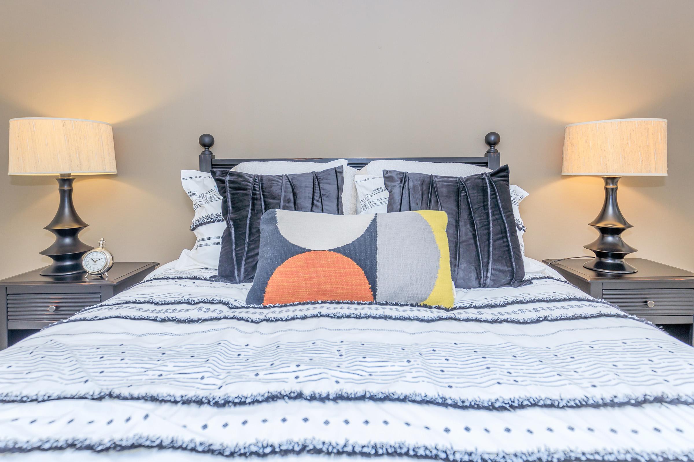 A neatly made bed with a cozy comforter featuring a striped pattern, accented by a decorative pillow with geometric designs. Two stylish bedside lamps with fabric shades illuminate the space, alongside a small clock on one nightstand against a warm, neutral wall.