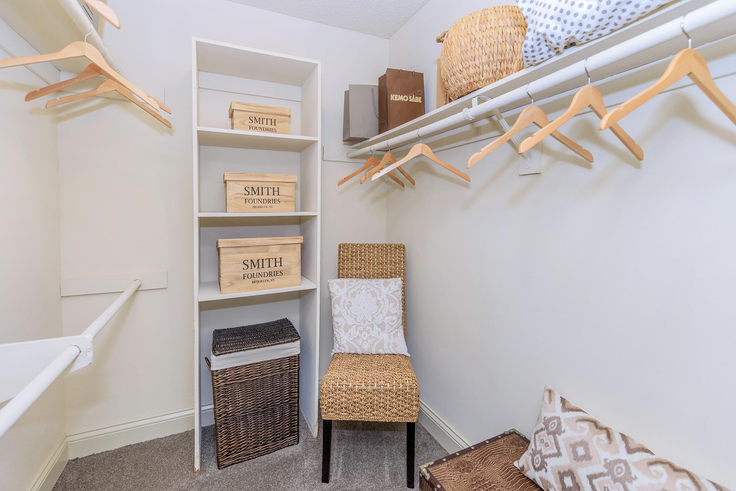 A well-organized closet featuring a shelf with wooden storage boxes labeled "SMITH FOUNDERS," empty wooden hangers, a woven chair, and soft storage bins on the floor, all against a light-colored wall and plush carpet.