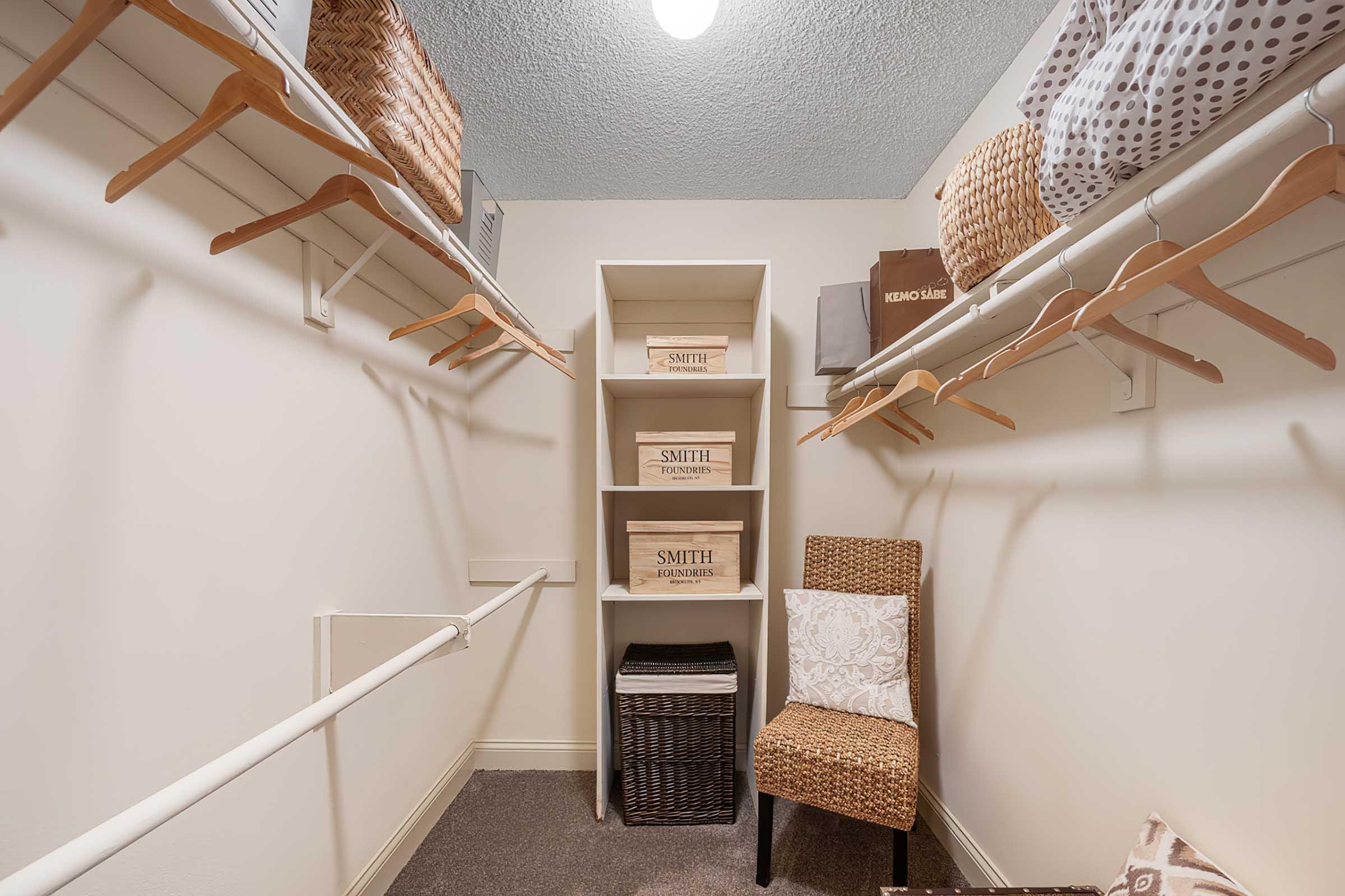 A well-organized closet featuring wooden hangers, wicker baskets, and storage boxes labeled "SMITH." A single chair with a cushion sits in the corner. The walls are painted in a light tone, and a ceiling light provides illumination, creating a clean and tidy space for storing clothes and accessories.