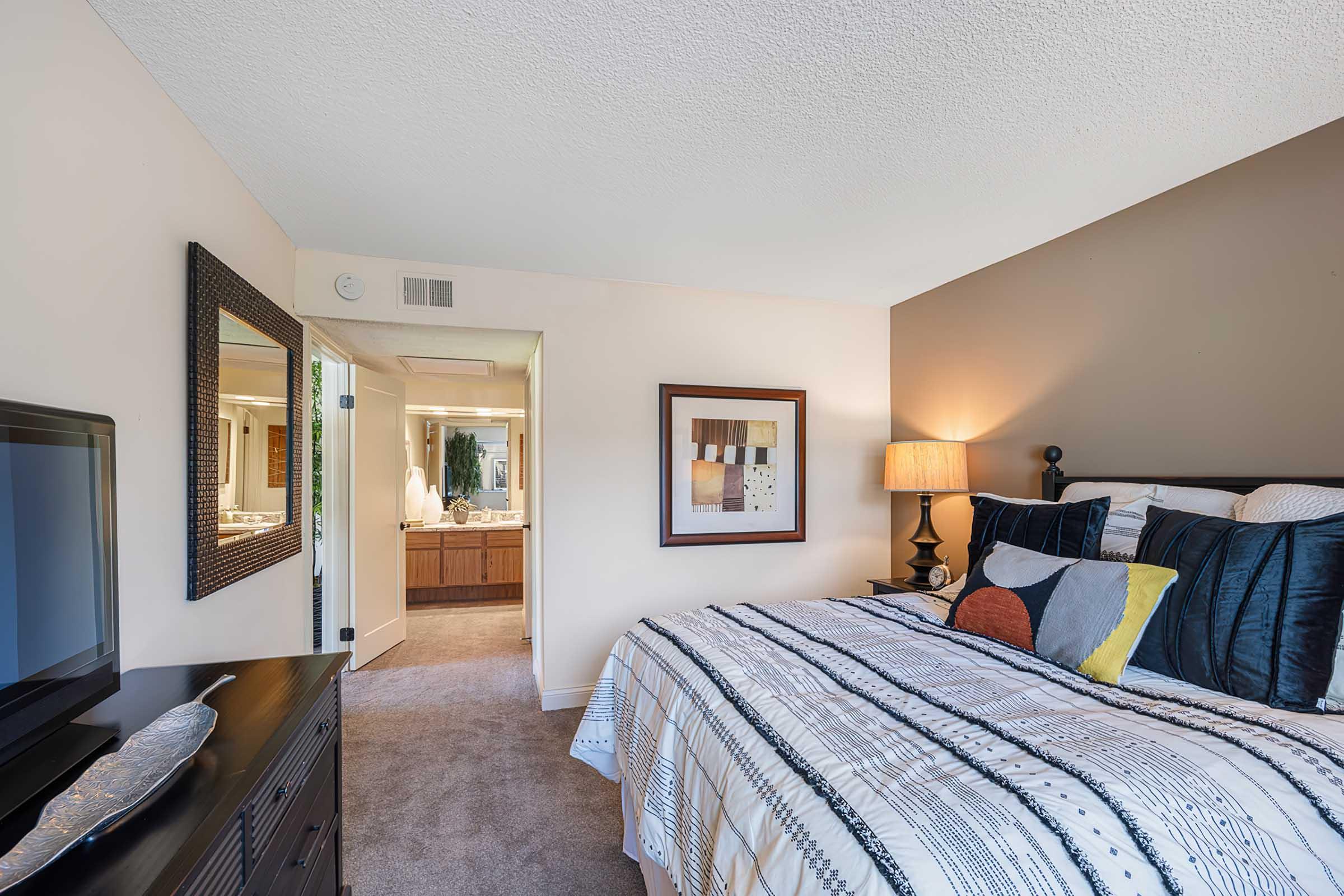 A cozy bedroom featuring a neatly made bed with decorative pillows, a bedside lamp, and a dresser with a television. A large mirror hangs on the wall, and an open doorway leads to a spacious bathroom area, illuminated by soft lighting. The walls have a warm, neutral color scheme.