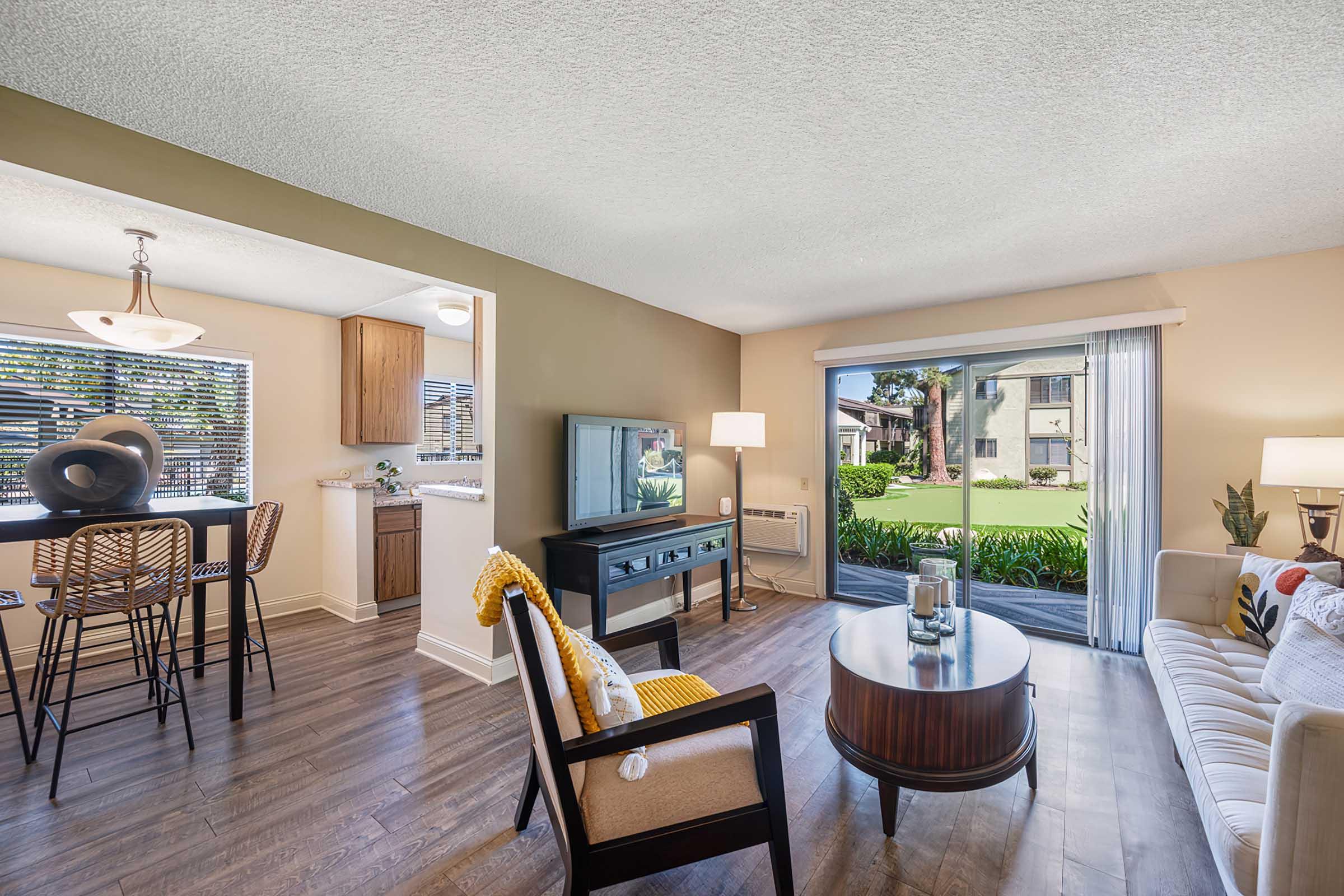 A modern living room with a cozy couch, a round coffee table, and a stylish chair. A television is on a console, and there is a lamp beside the couch. Large windows offer a view of greenery outside. A dining area with bar stools is adjacent, featuring a light fixture overhead and stylish cabinetry in the background.