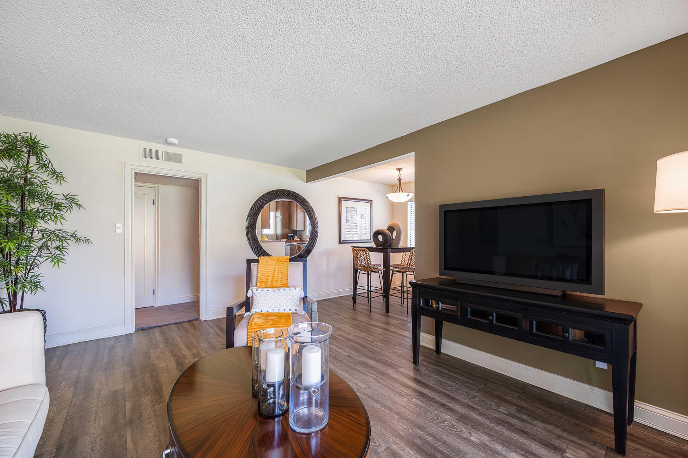 A modern living room featuring a neutral color palette with a comfortable chair, a round mirror, and a coffee table with candles. A large TV is mounted on the wall, and a dining area is visible in the background, creating an inviting and stylish atmosphere.