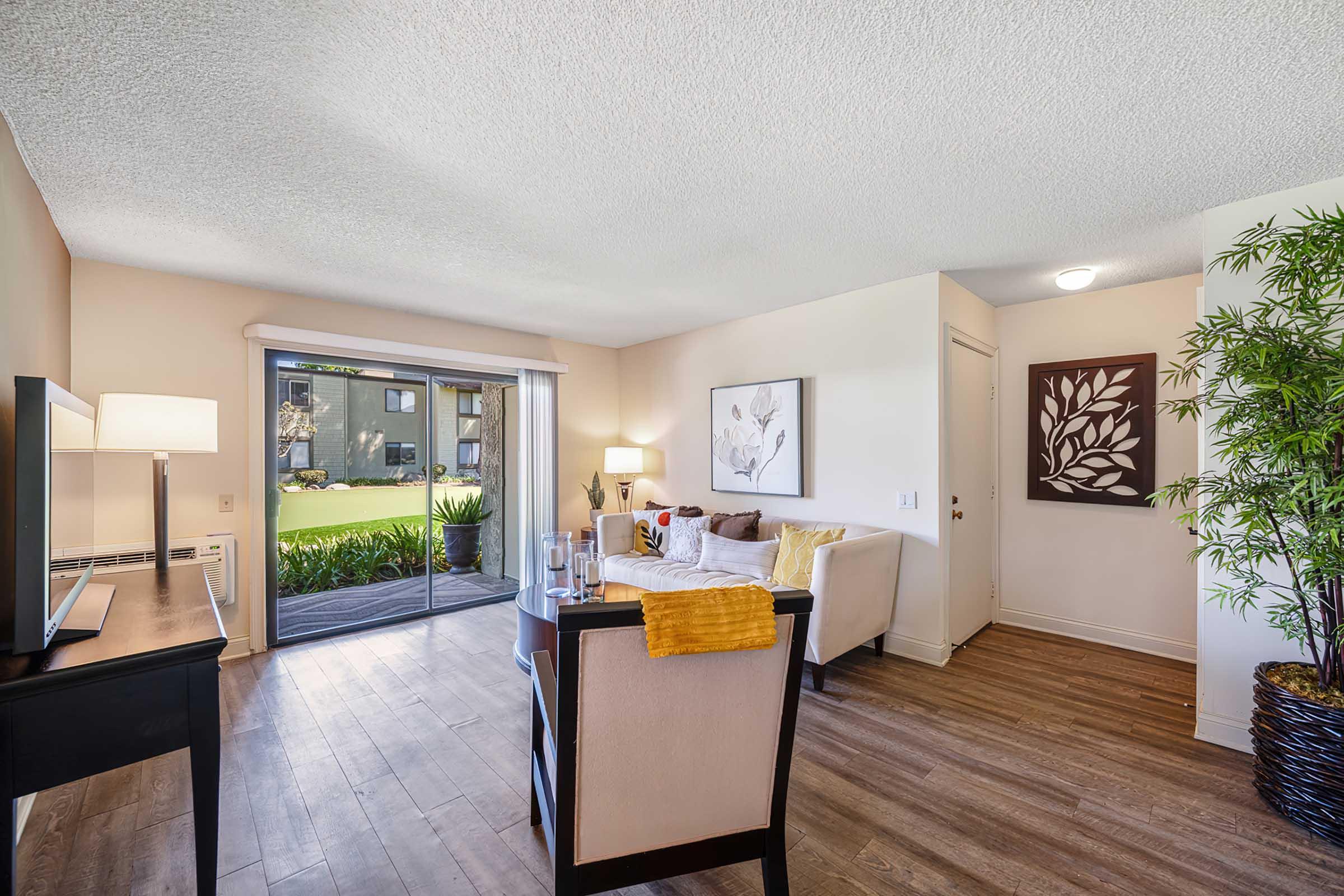 Bright and inviting living space with wood flooring, large windows overlooking a green exterior. Features a comfortable sofa, a decorative armchair, a dining table, and a lamp. Wall art adds a touch of style, while potted plants provide a natural element to the decor.