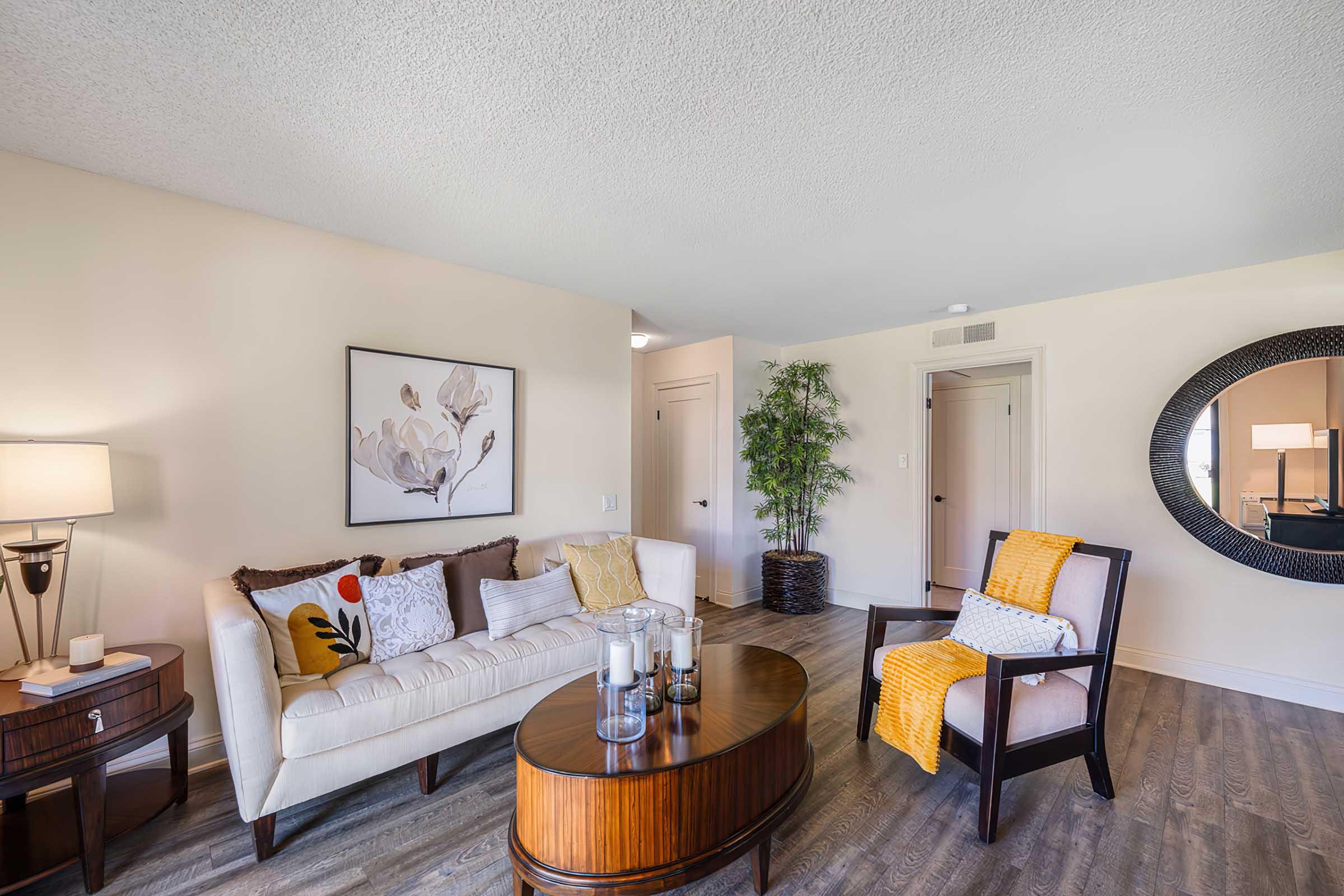 A modern living room featuring a white couch adorned with decorative pillows, a round wooden coffee table with candles, and a yellow throw blanket on a wooden armchair. There's a large mirror on the wall, a potted plant in the corner, and natural light streaming in from the doorway.