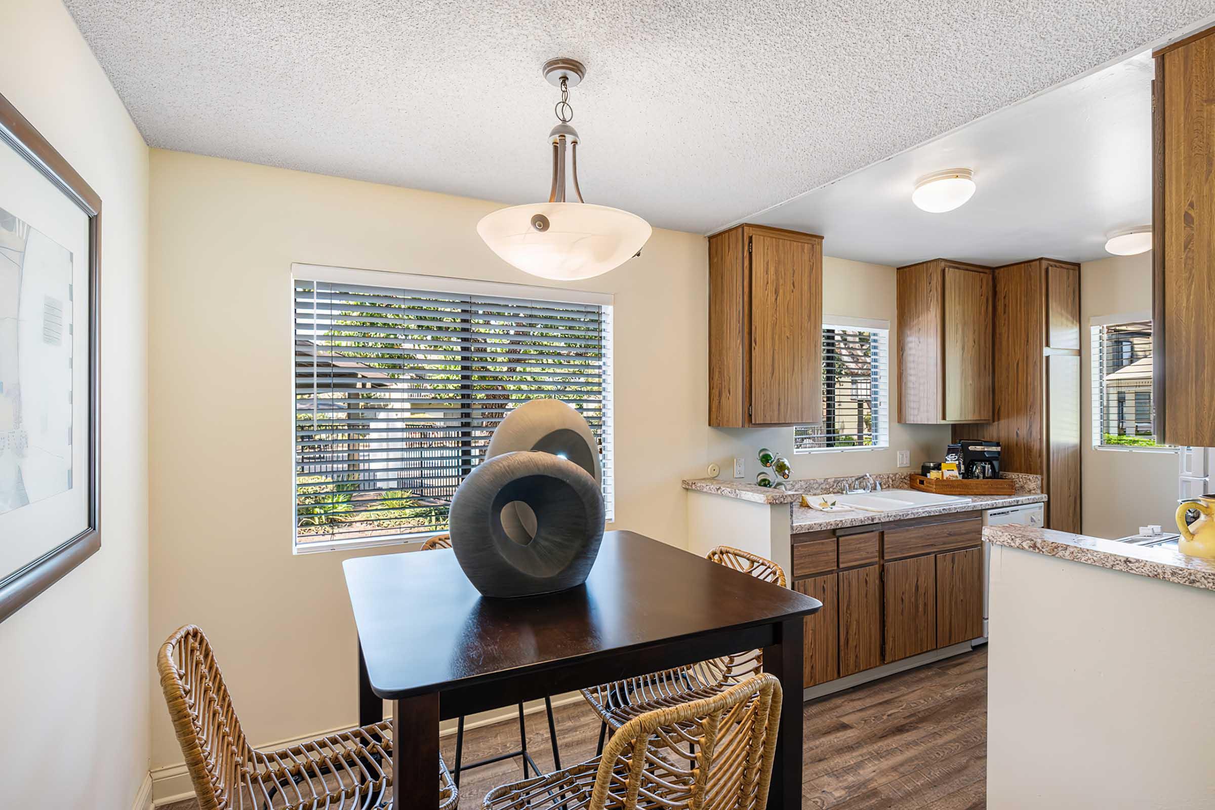 A modern kitchen with wooden cabinets and a small dining table set for two. The space has a large window with blinds allowing natural light, and decorative items on the counter. The decor features neutral colors, creating a warm and inviting atmosphere.