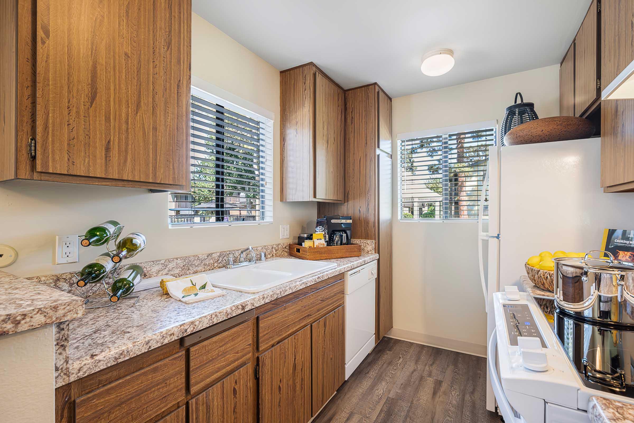 A modern kitchen featuring wooden cabinets, a granite countertop, a white sink, and a large window with blinds. There is a white refrigerator, a stovetop with pots, and a coffee machine. A few bottles of wine are displayed beside the sink, and fresh lemons are placed on the countertop.