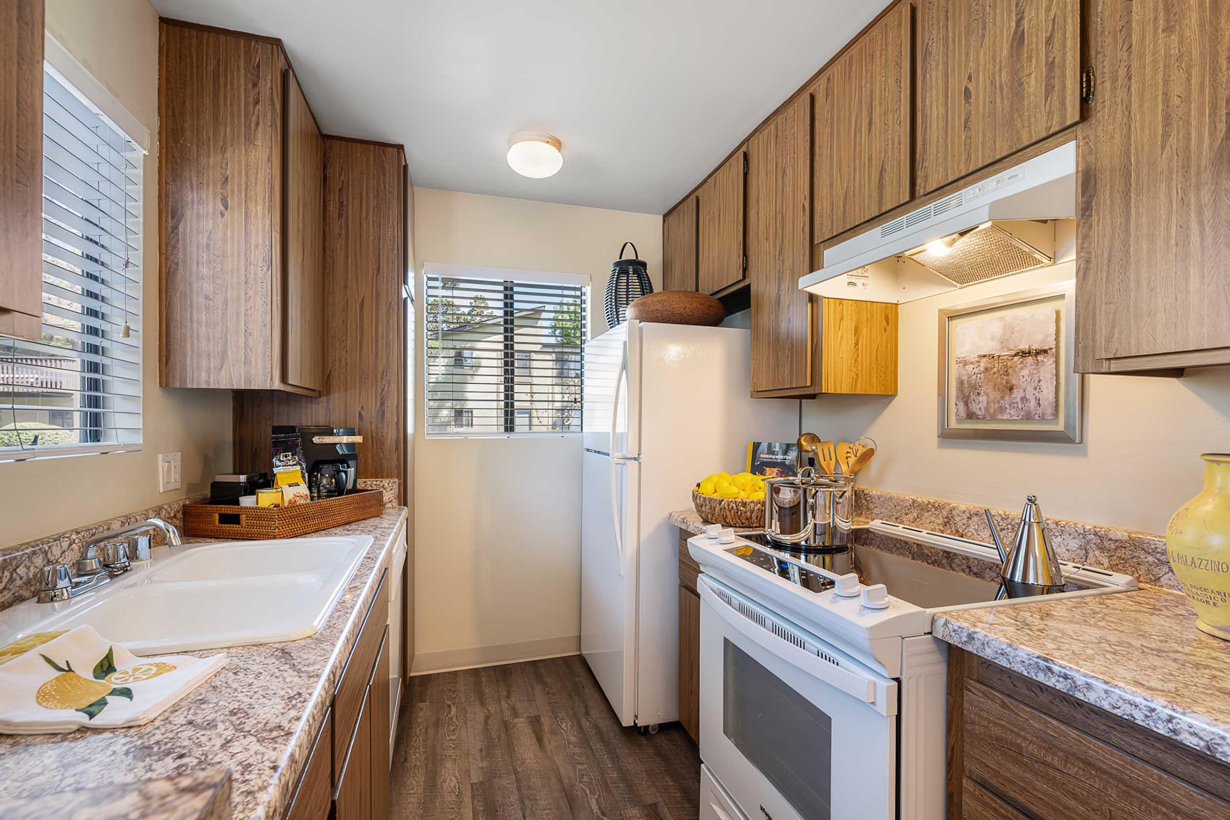 Modern kitchen with wooden cabinetry and granite countertops. Features a white stove, refrigerator, and a coffee maker, along with a bowl of lemons and kitchen utensils. There are windows providing natural light and a decorative wall art piece. The space is organized and clean, showcasing a cozy cooking environment.