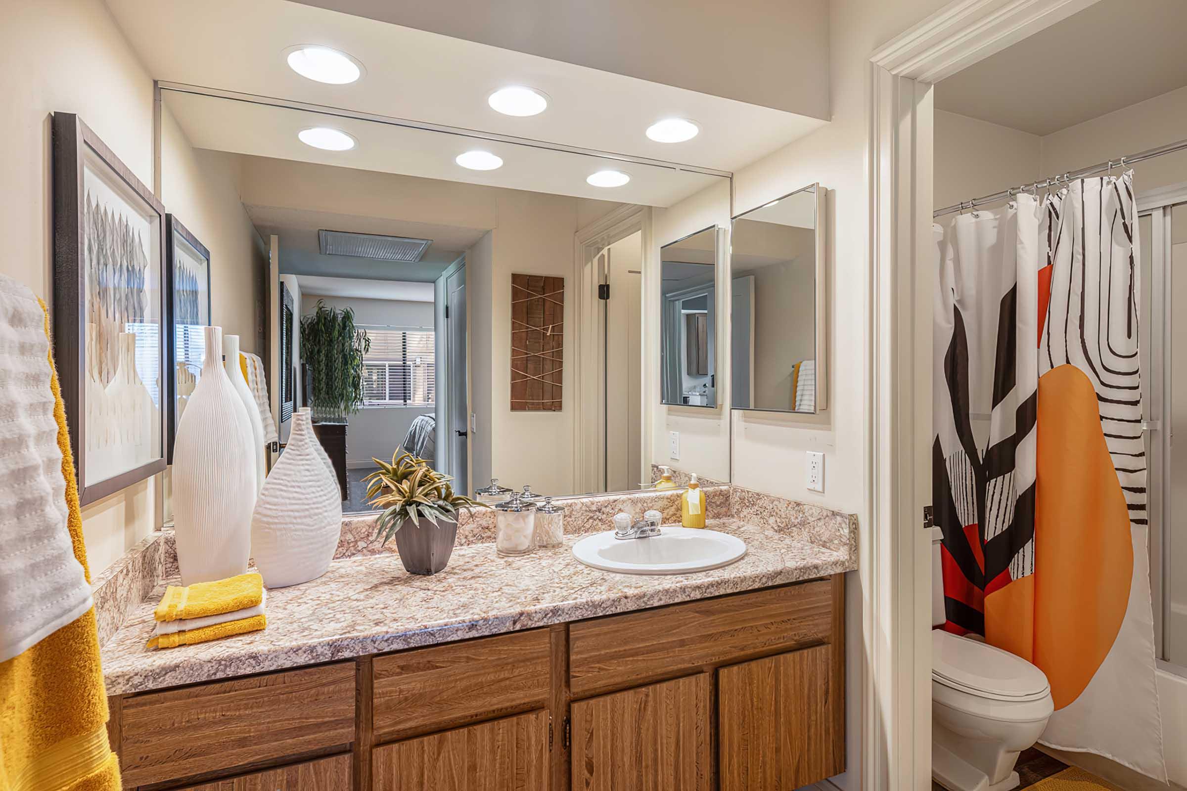 A modern bathroom featuring a countertop with a decorative vase and a small plant, a large mirror reflecting the space, and bright lighting. The walls are neutral-colored, with a colorful shower curtain visible, and towels neatly arranged nearby.