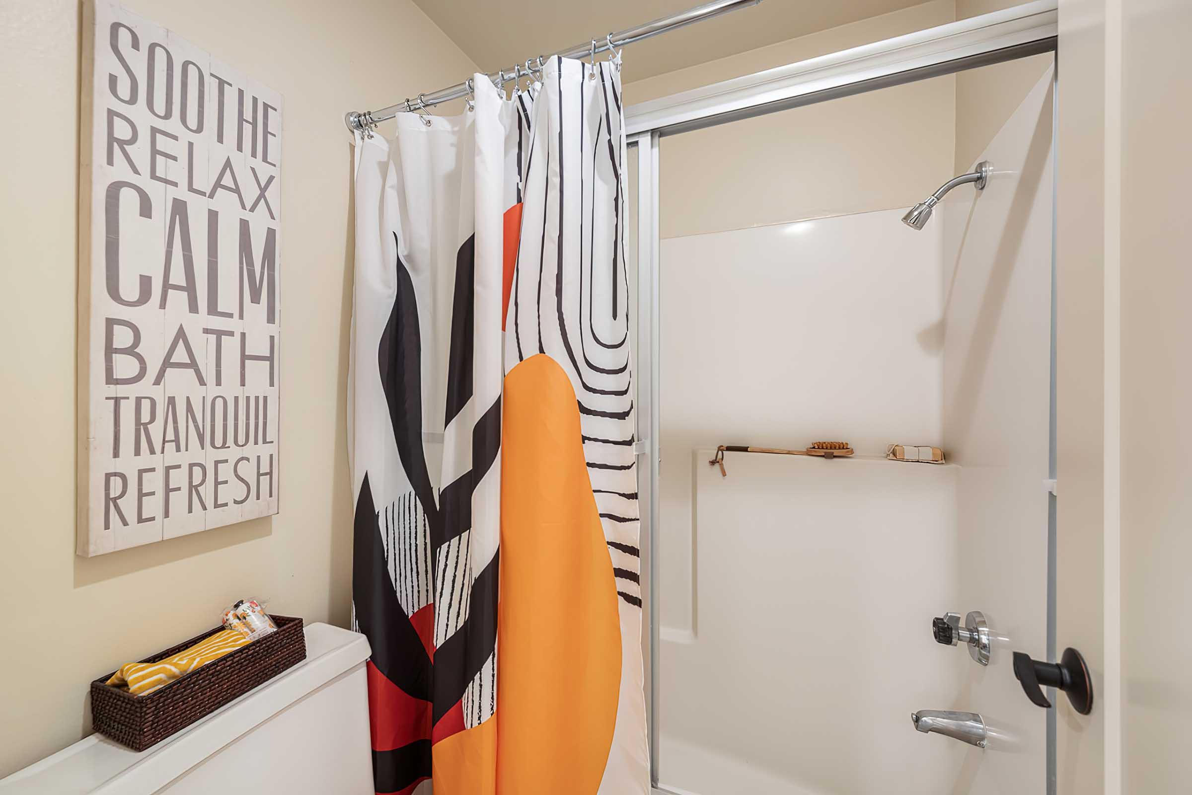 A bathroom shower with a colorful curtain featuring abstract patterns. A wall-mounted shelf holds bath accessories. A text-art print on the wall encourages relaxation and tranquility. The overall atmosphere is calm and inviting.