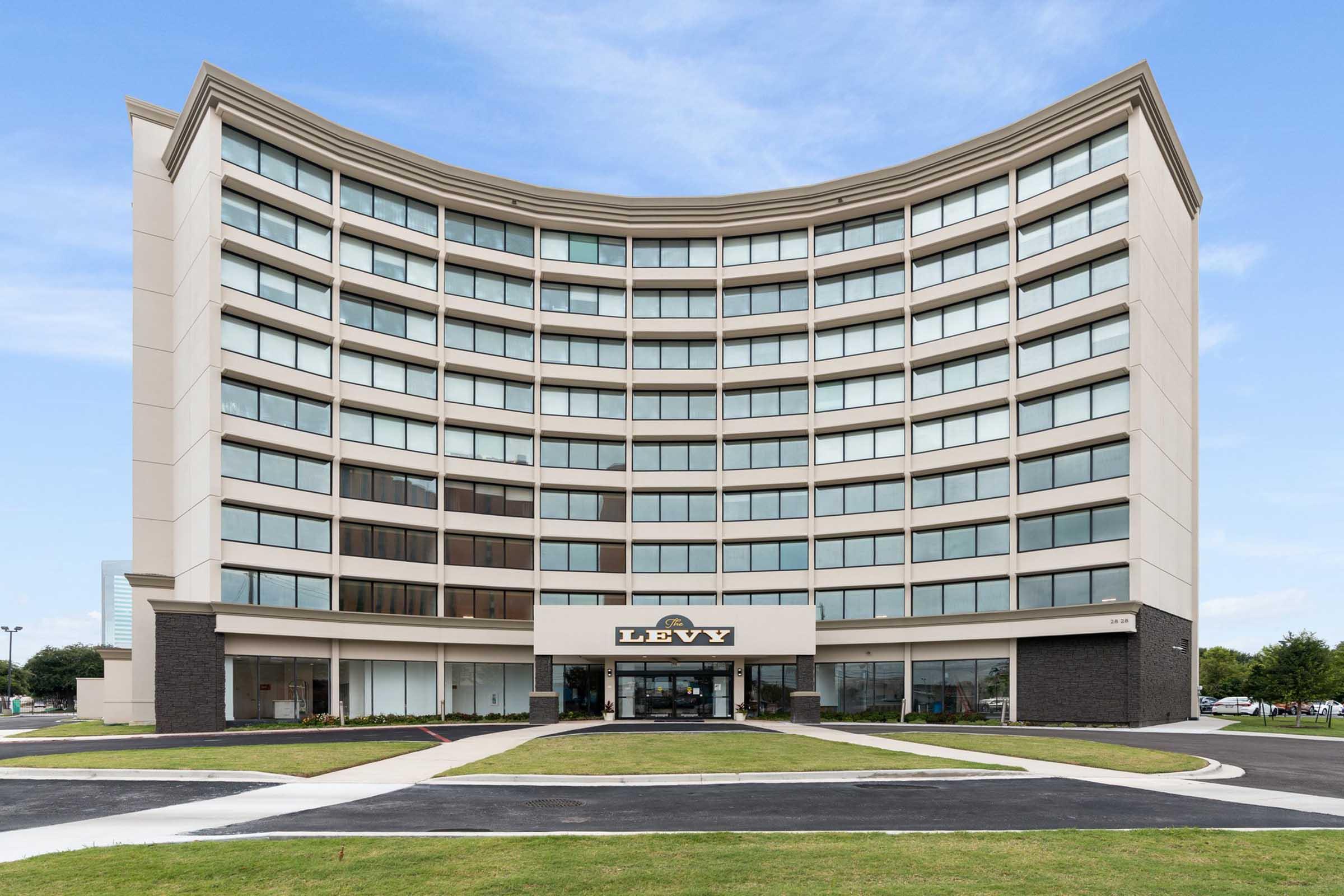 A modern, curved building showcasing the name "LEVY" prominently at the entrance. The structure has multiple large windows and a contemporary design, set against a clear blue sky. The surrounding area features neatly maintained grass and paved pathways.