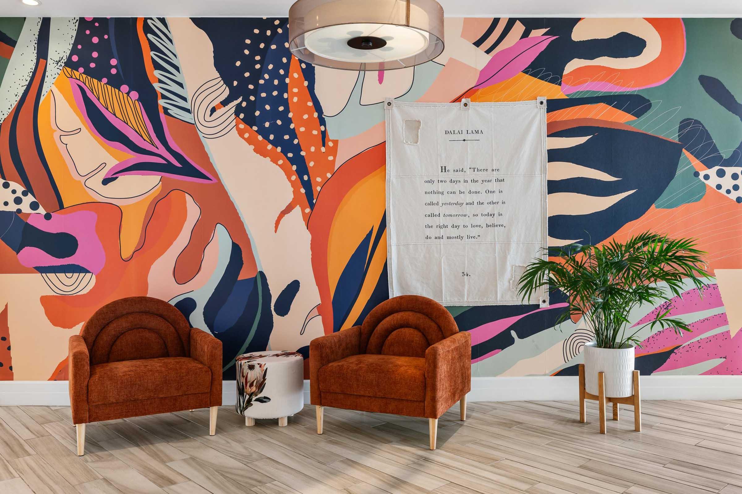A modern interior featuring two orange armchairs with curved backs in front of a vibrant wall mural of abstract shapes and colors. A white textile wall hanging with text is displayed beside the chairs, and a small potted plant adds greenery to the scene. The floor is light wood.