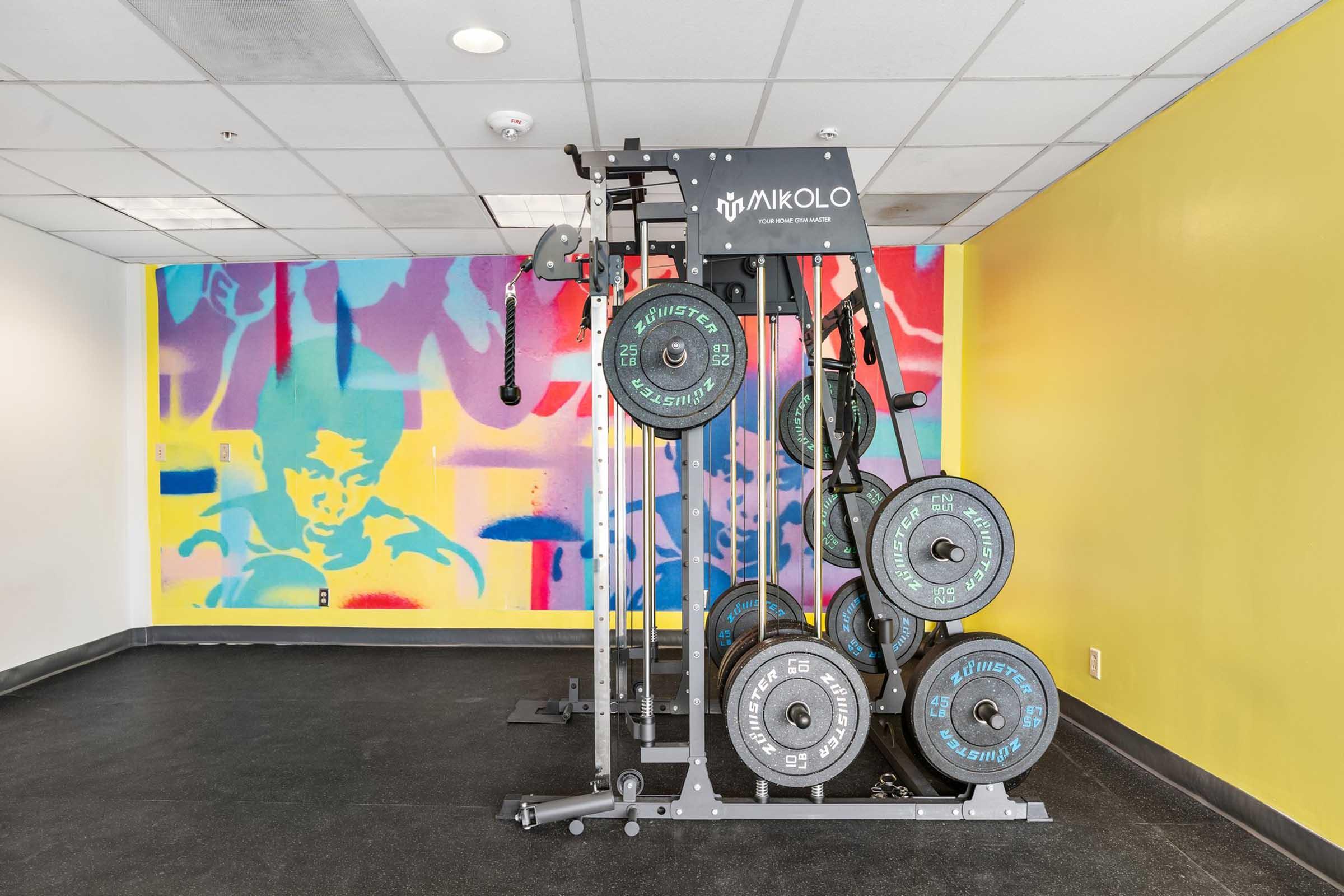 A strength training machine with weight plates attached, positioned against a colorful mural featuring abstract figures and shapes. The room has a modern design with bright yellow walls and a dark floor, creating an energetic atmosphere suitable for workouts.
