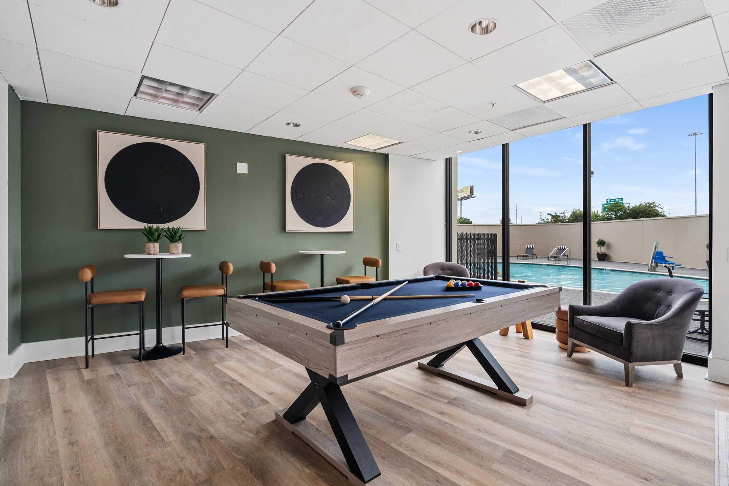 A modern lounge area featuring a billiards table, two round tables with chairs, and a comfortable armchair. Large windows provide natural light and a view of an outdoor pool area. The walls are painted in a green hue with two circular artworks displayed. The flooring is light wood.