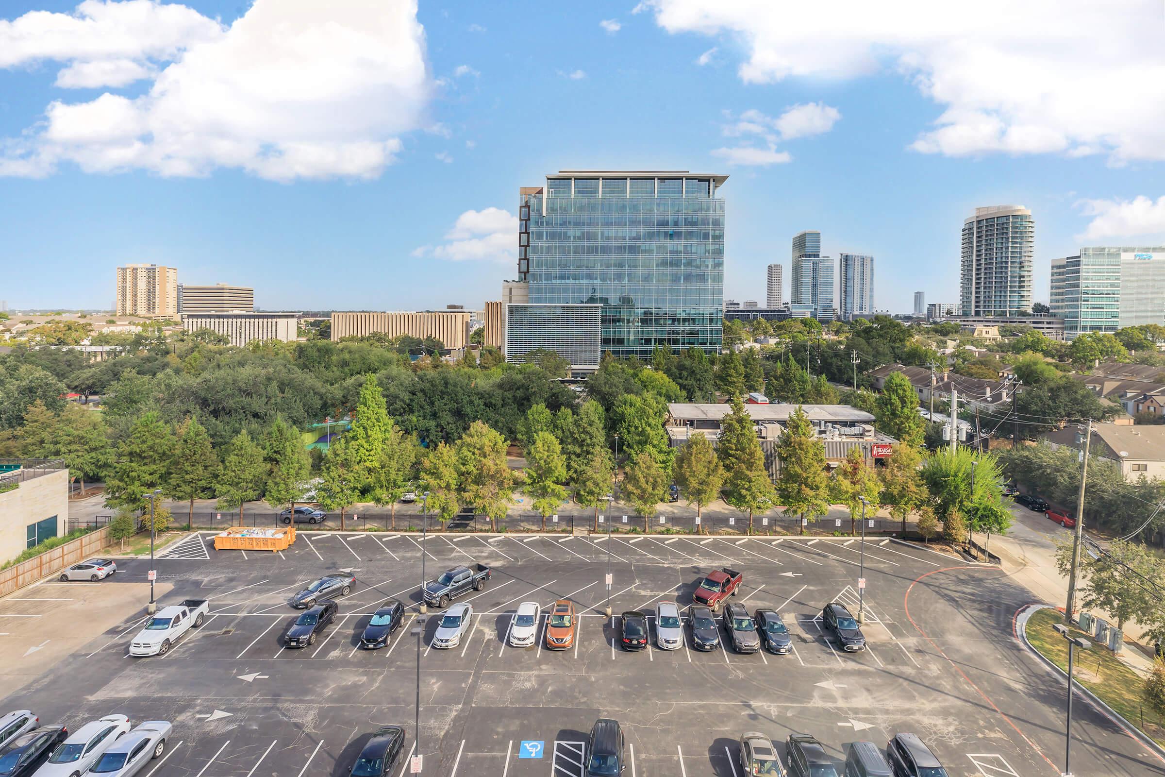 A view of a city skyline featuring modern buildings and a green landscape. In the foreground, a parking lot is visible with several parked cars. The background includes high-rise structures and a clear blue sky with scattered clouds.