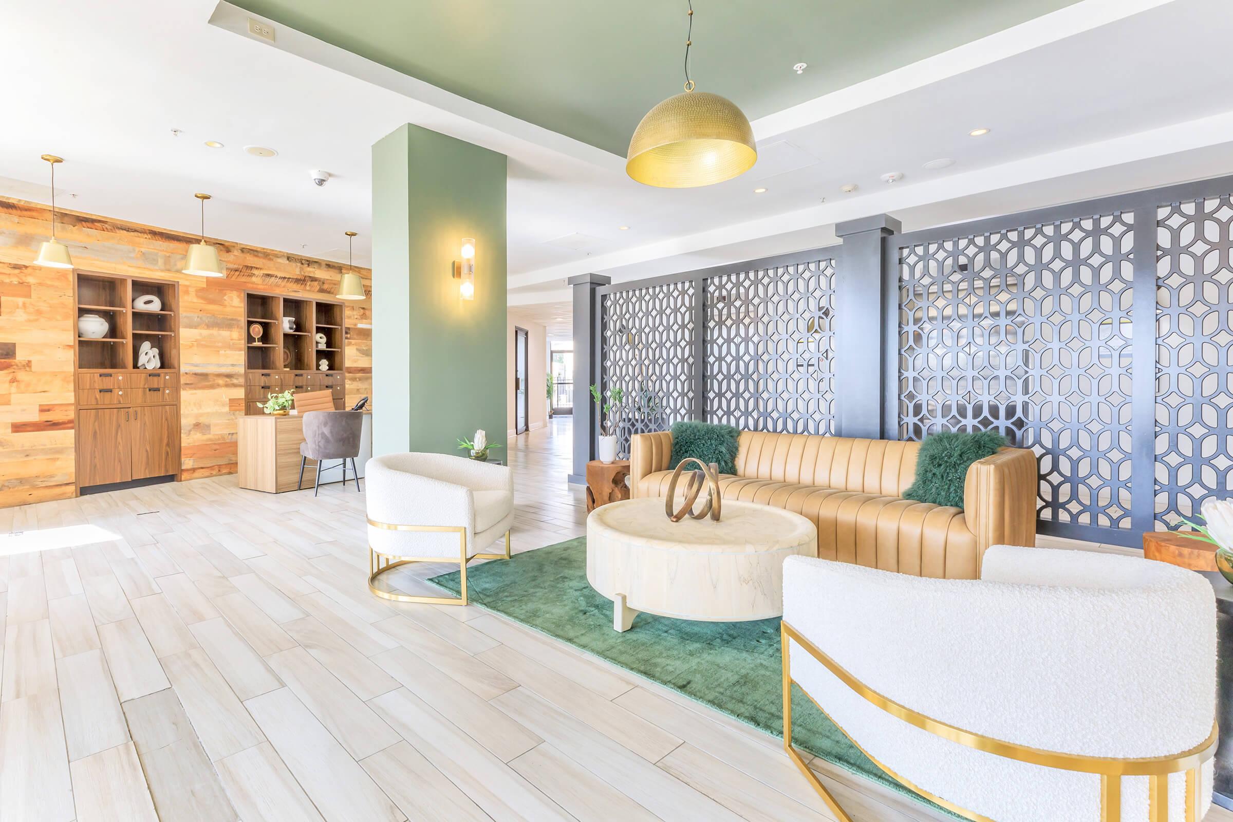 A modern lounge area featuring a combination of a beige leather sofa, round coffee table, and plush white chairs. The space is accented with green plants and décor, complemented by wooden panels and patterned dividers. Natural light streams in, creating a welcoming atmosphere.
