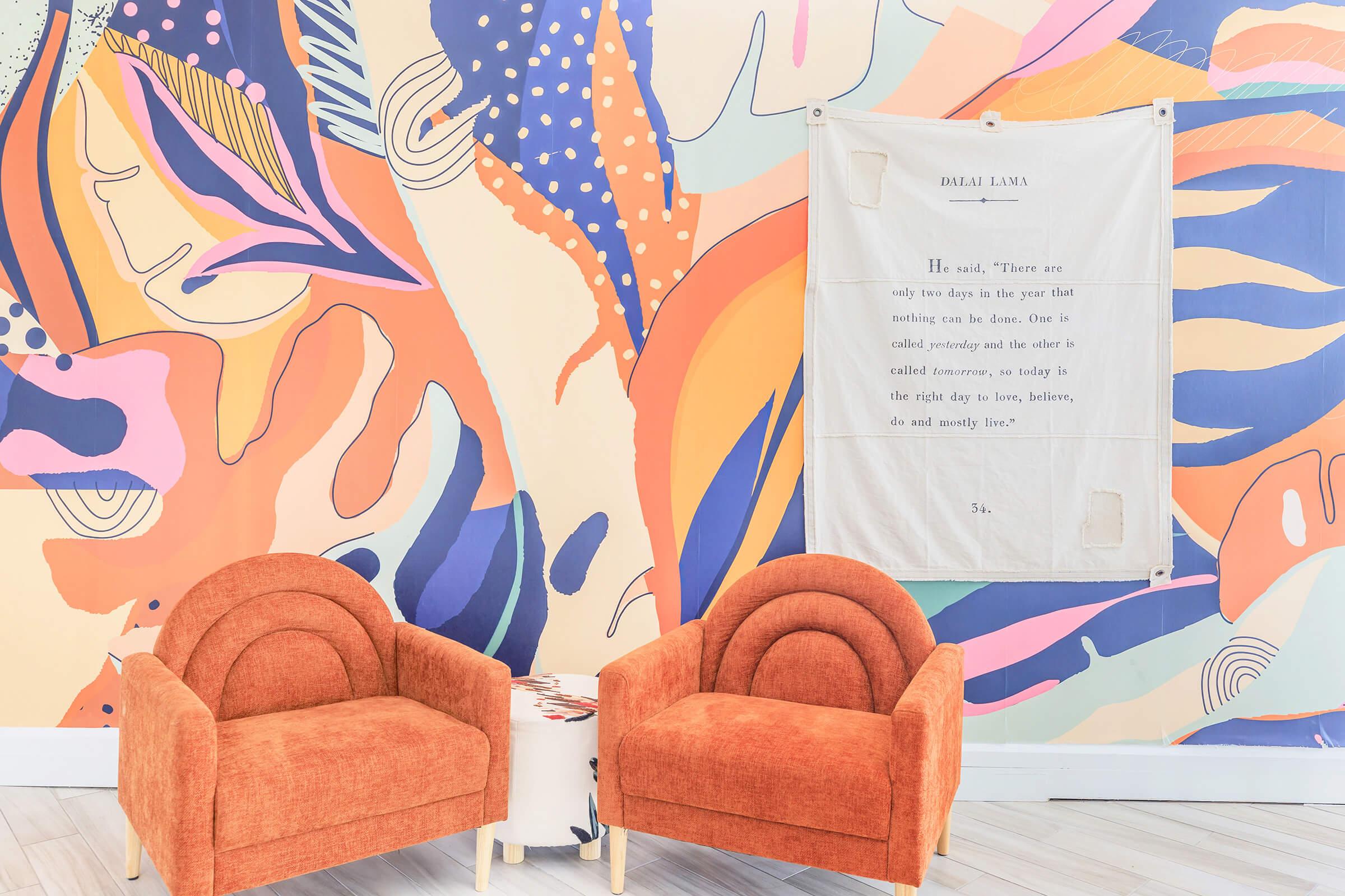 A colorful abstract mural featuring vibrant shapes and patterns serves as a backdrop to two orange, curved armchairs placed in front of a small white table. A fabric wall hanging with a quote attributed to the Dalai Lama is displayed nearby, adding a spiritual touch to the modern, inviting space.