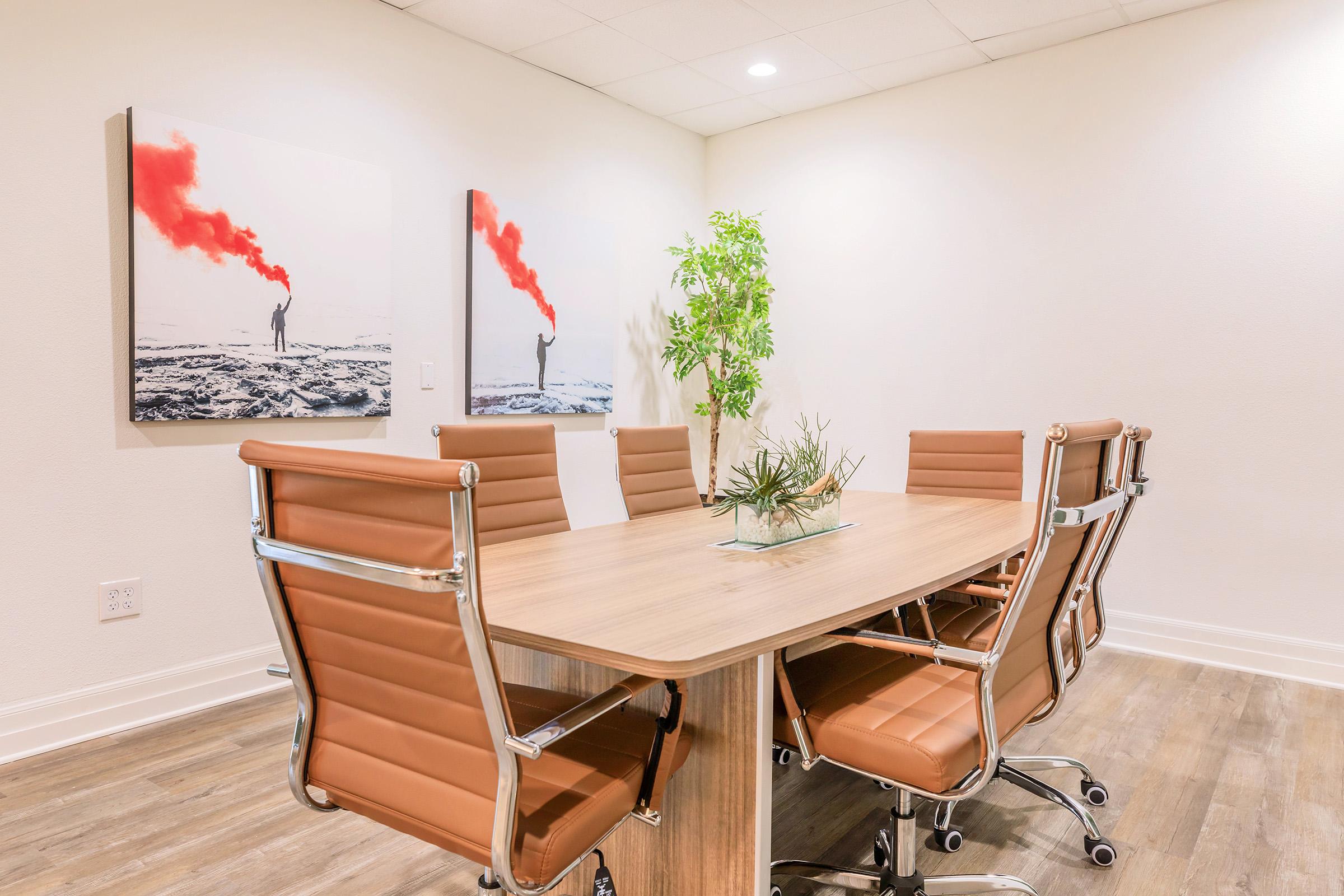 A modern conference room featuring a sleek wooden table surrounded by stylish brown leather chairs. Two abstract art pieces with red smoke effects are mounted on the wall, and a small green plant is placed on the table, adding a touch of nature to the contemporary decor.