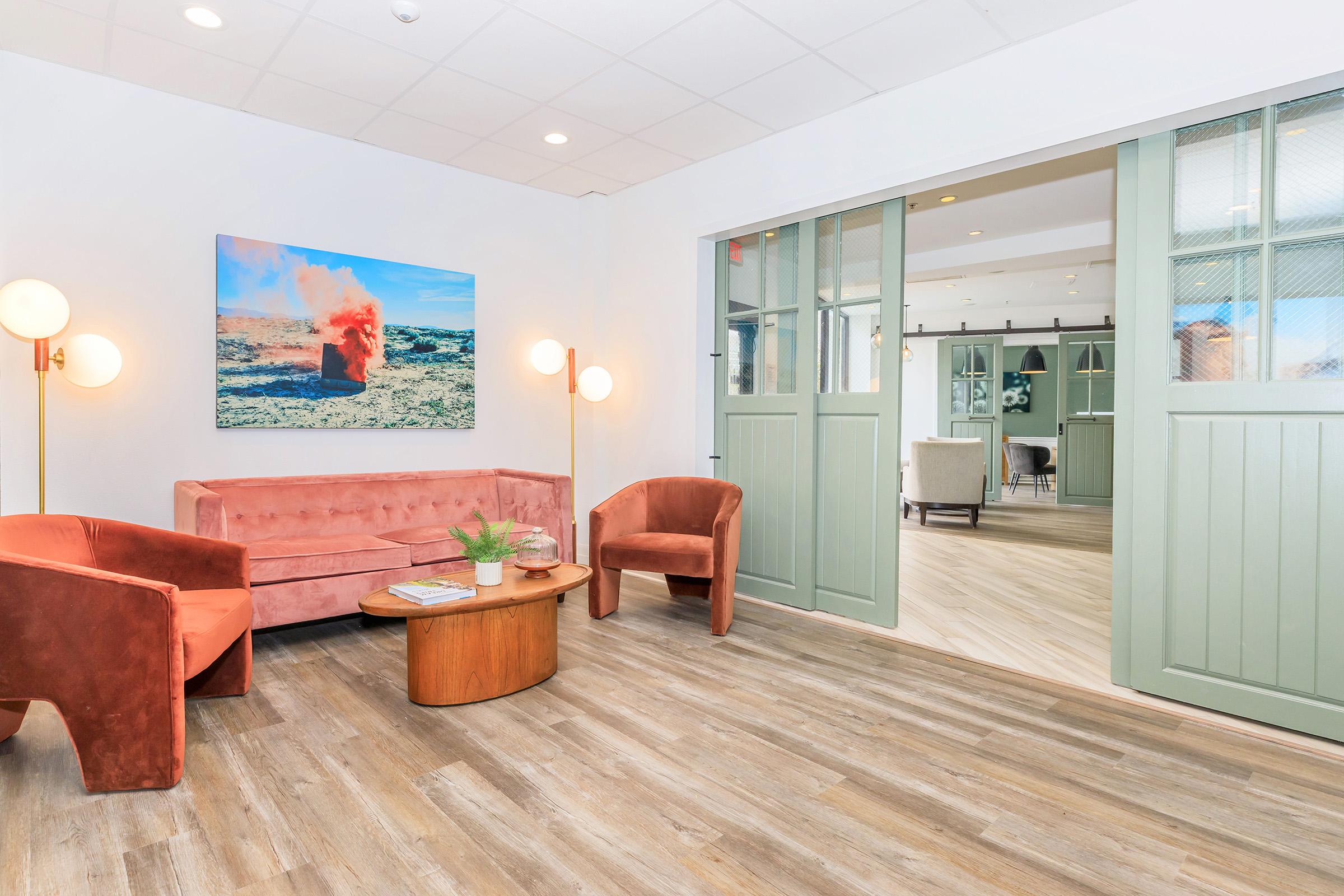 A modern interior space featuring a pink velvet sofa and two orange armchairs around a wooden coffee table. The room has a light wooden floor and is adorned with a colorful abstract painting on the wall. A set of green sliding doors leads to another area in the background, creating an inviting atmosphere.