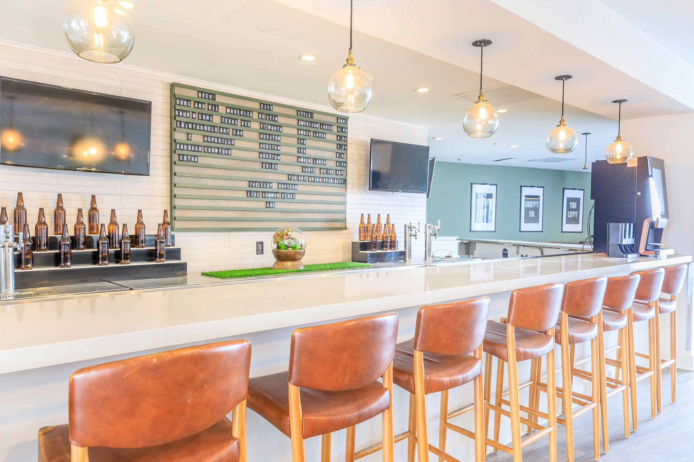 A modern bar with a sleek countertop and wooden stools, featuring illuminated hanging light fixtures. Behind the bar, there is a wall displaying various beer options and two flat-screen TVs. The atmosphere is bright and inviting, suitable for social gatherings or relaxation.