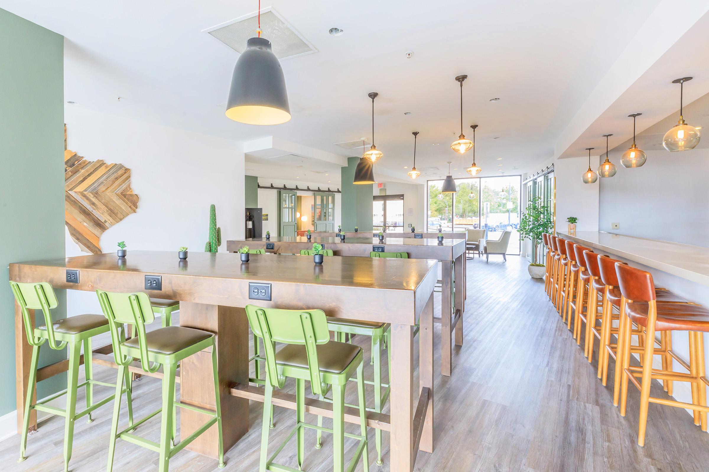 Interior of a bright, modern dining space featuring wooden tables with green bar stools, pendant lighting, and large windows. There are plants and decorative elements, creating a welcoming atmosphere suitable for social gatherings or dining.