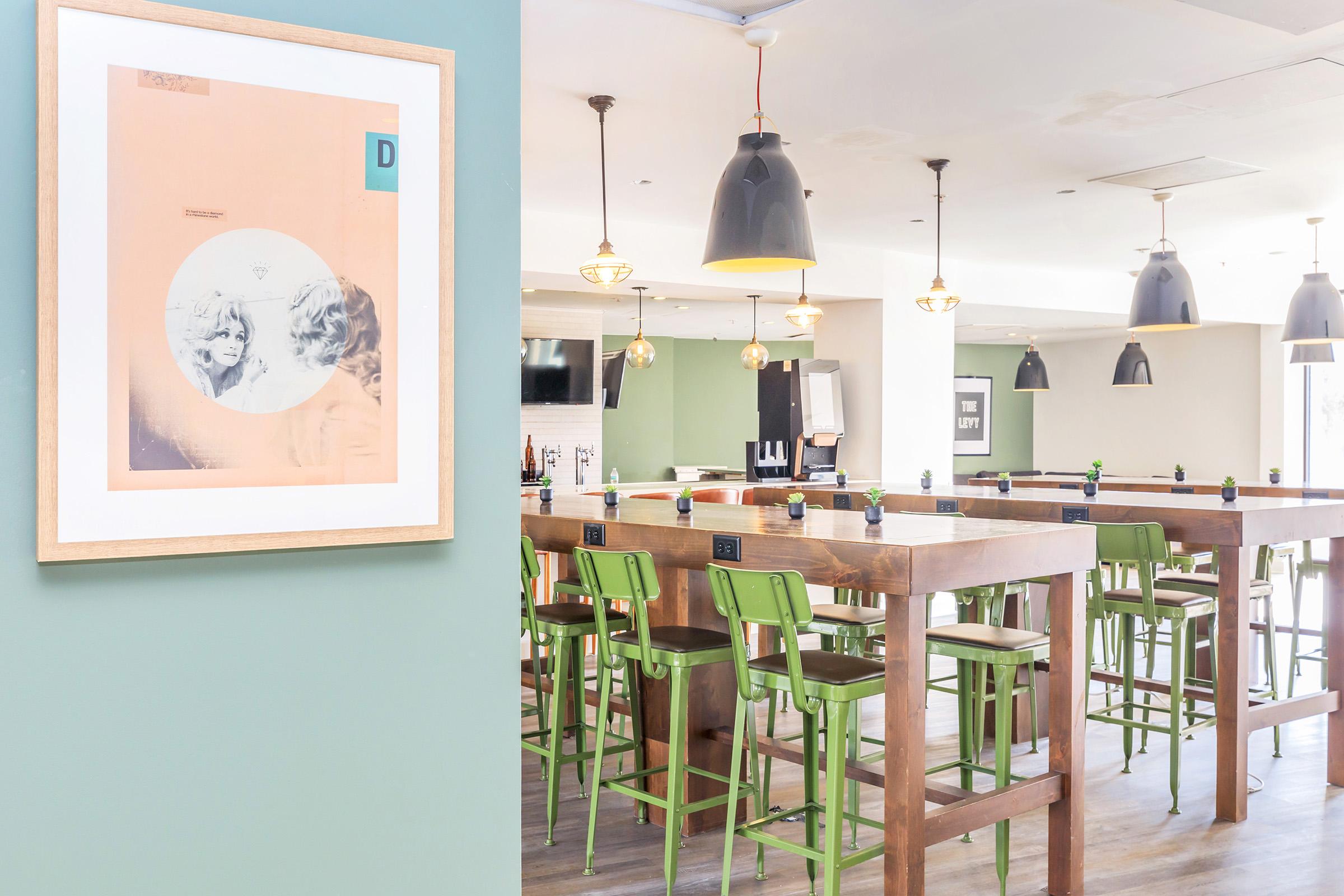 A modern interior of a restaurant or bar featuring a wooden table with green chairs, pendant lighting overhead, and decorative framed artwork on the wall. The space has a bright and inviting atmosphere with a mix of light colors and textures.
