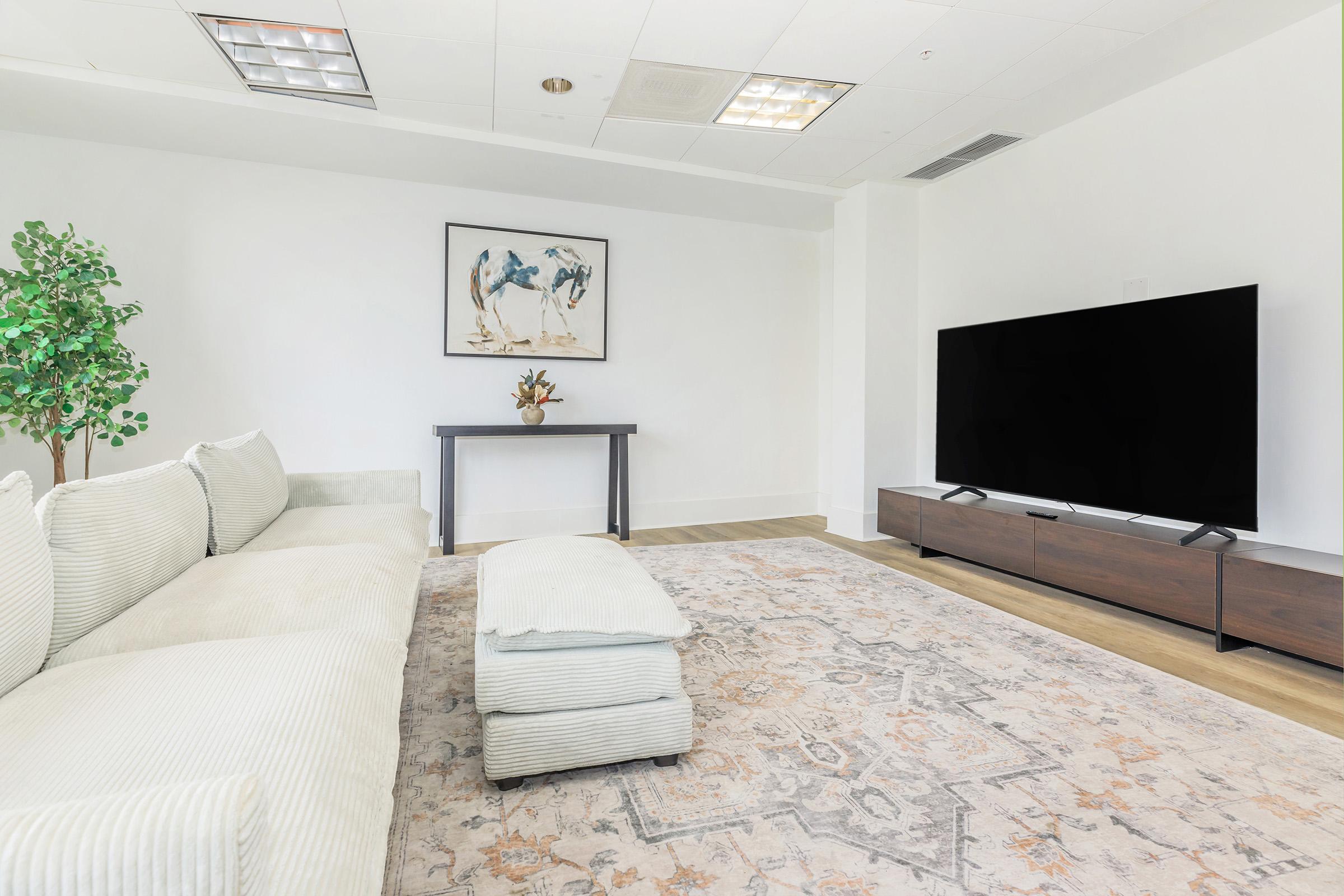 A modern living room featuring a light-colored sofa, a small ottoman, and a dark wooden TV stand with a large flat-screen TV. There's a wall-mounted artwork above a wooden table with a vase of flowers. A potted plant adds a touch of greenery to the bright, spacious area with light hardwood flooring.