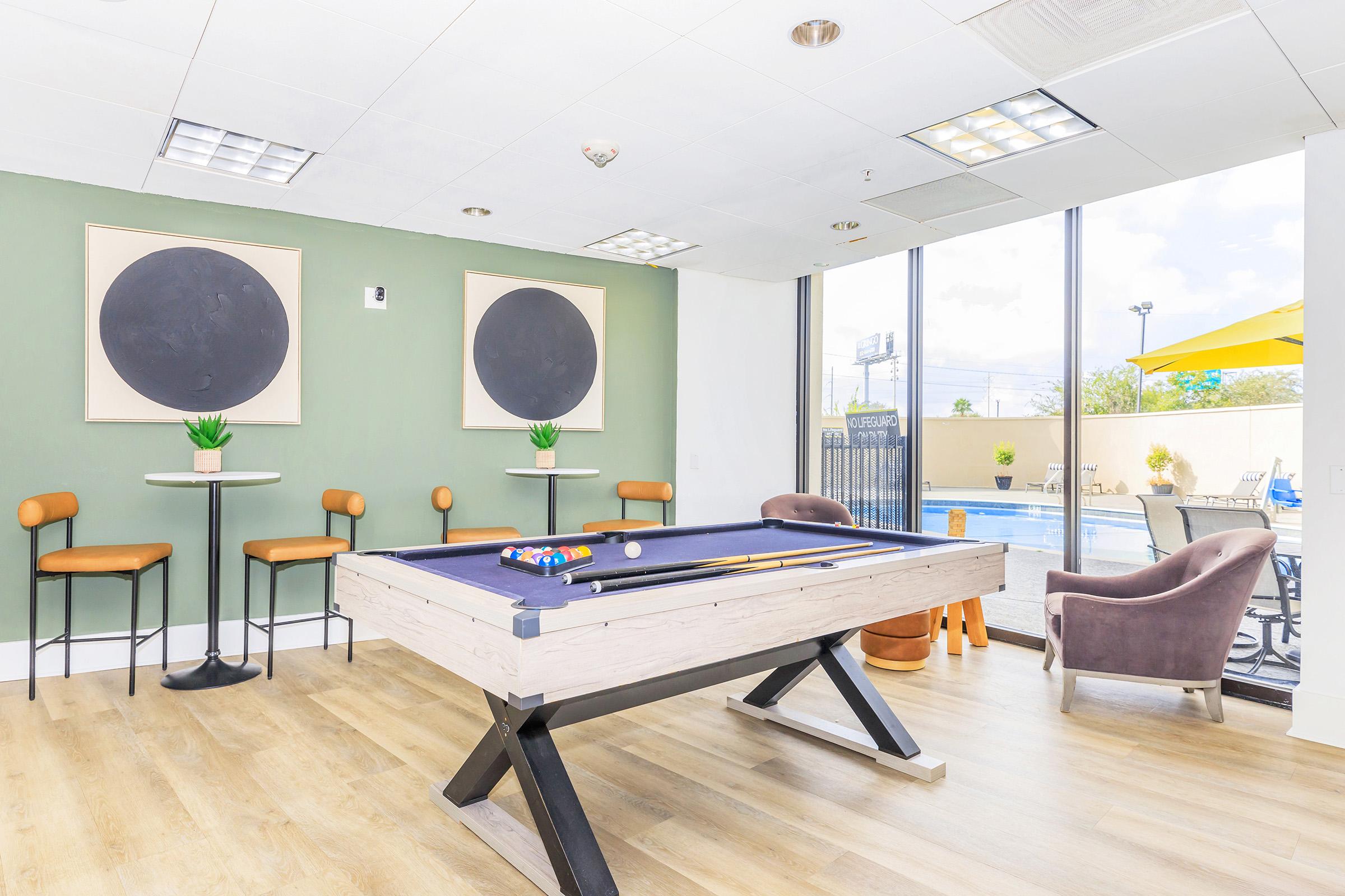 A bright room featuring a pool table in the center, surrounded by tall chairs and small tables. Two large circular art pieces adorn the green wall, and large windows offer a view of a pool area outside, with outdoor seating and a bright sky visible.