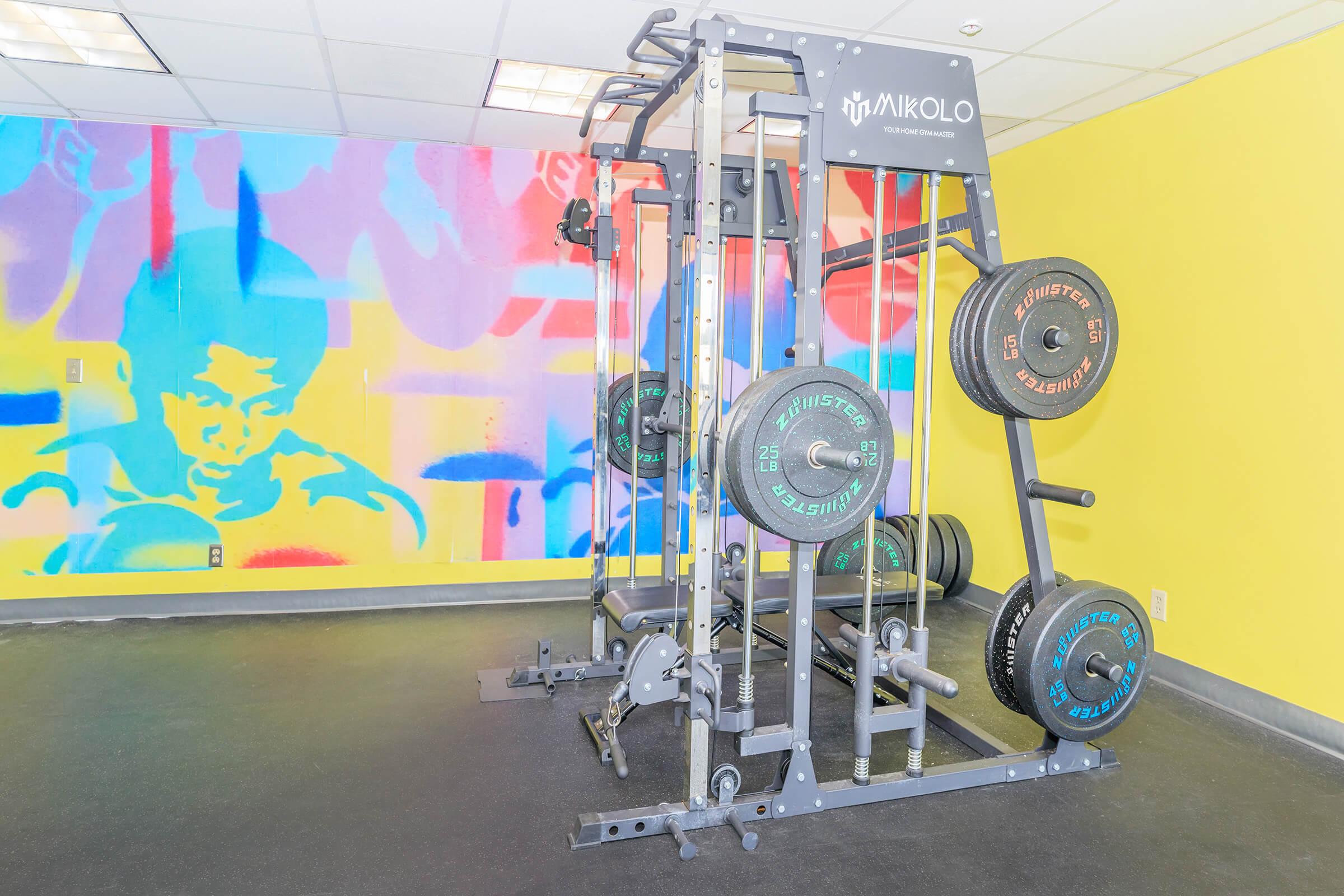 A modern gym setup featuring a multi-functional weight training machine with various weights attached. The background features a vibrant, colorful mural depicting athletic figures, creating a lively atmosphere in the fitness space. The flooring is dark, emphasizing the gym equipment.