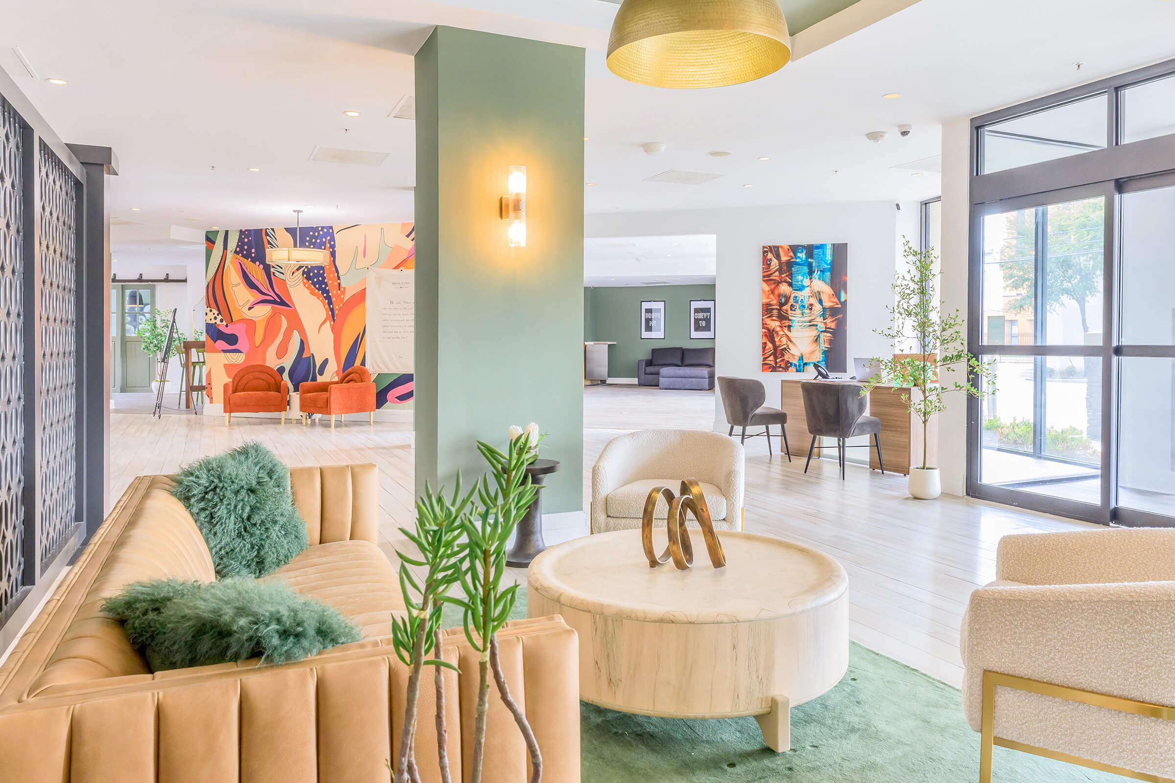 A modern lobby area with stylish furnishings, including a beige sofa with green cushions, a round marble coffee table, and various seating options. Colorful wall art adds vibrancy to the space, complemented by potted plants and large windows allowing natural light. The decor is contemporary and inviting.