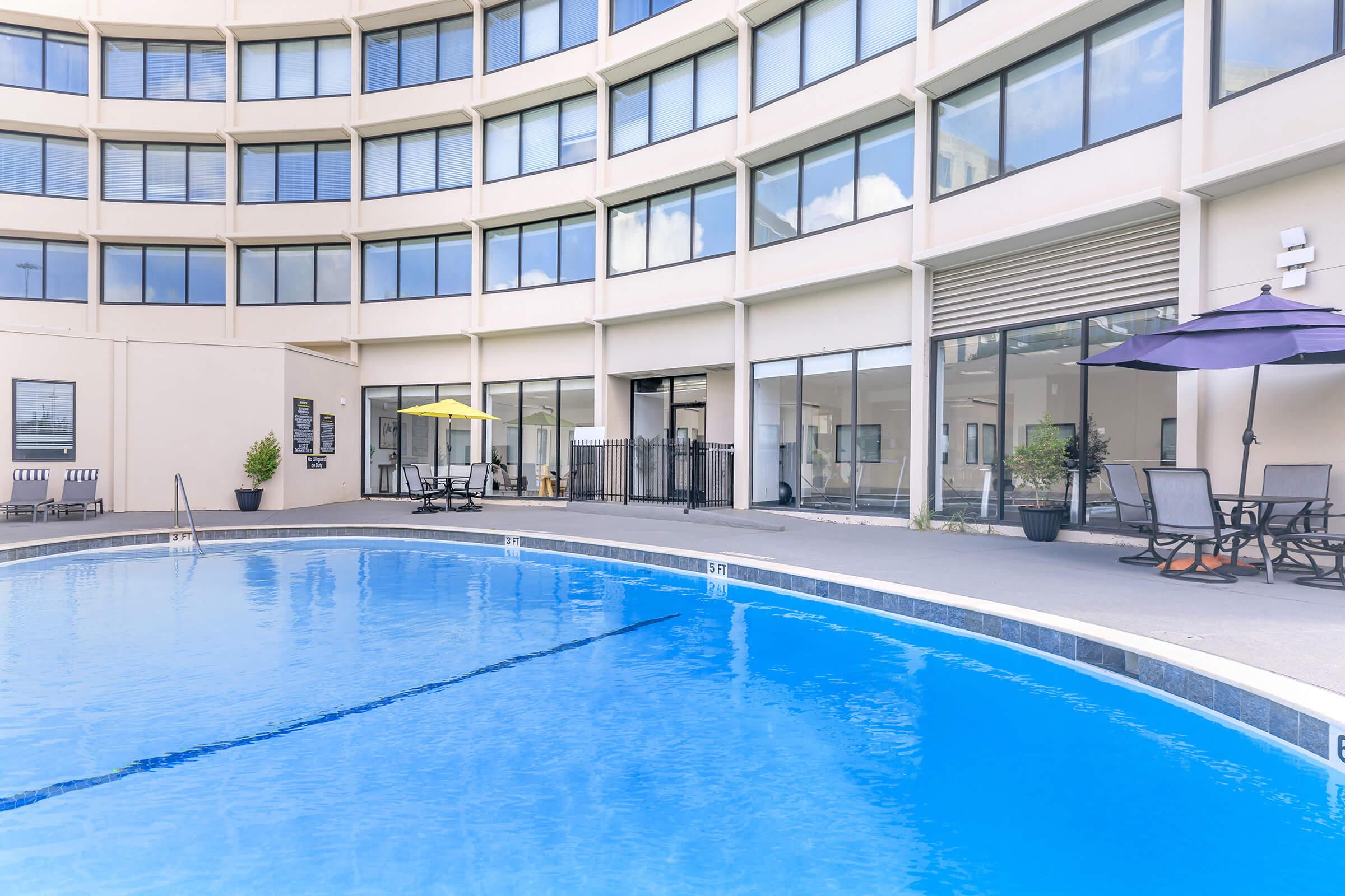 A clear blue swimming pool surrounded by a patio area with lounge chairs. The building features large windows and an outdoor seating area with yellow and purple umbrellas. The scene is bright and inviting, ideal for relaxation or leisure activities.