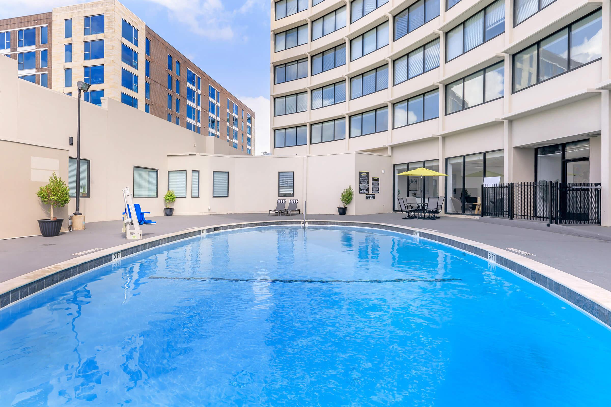 A clear blue swimming pool surrounded by a modern building with large windows. There are lounge chairs positioned near the pool, and a small green umbrella provides shade. The area is well-lit, with a clean and inviting atmosphere, ideal for relaxation and leisure.
