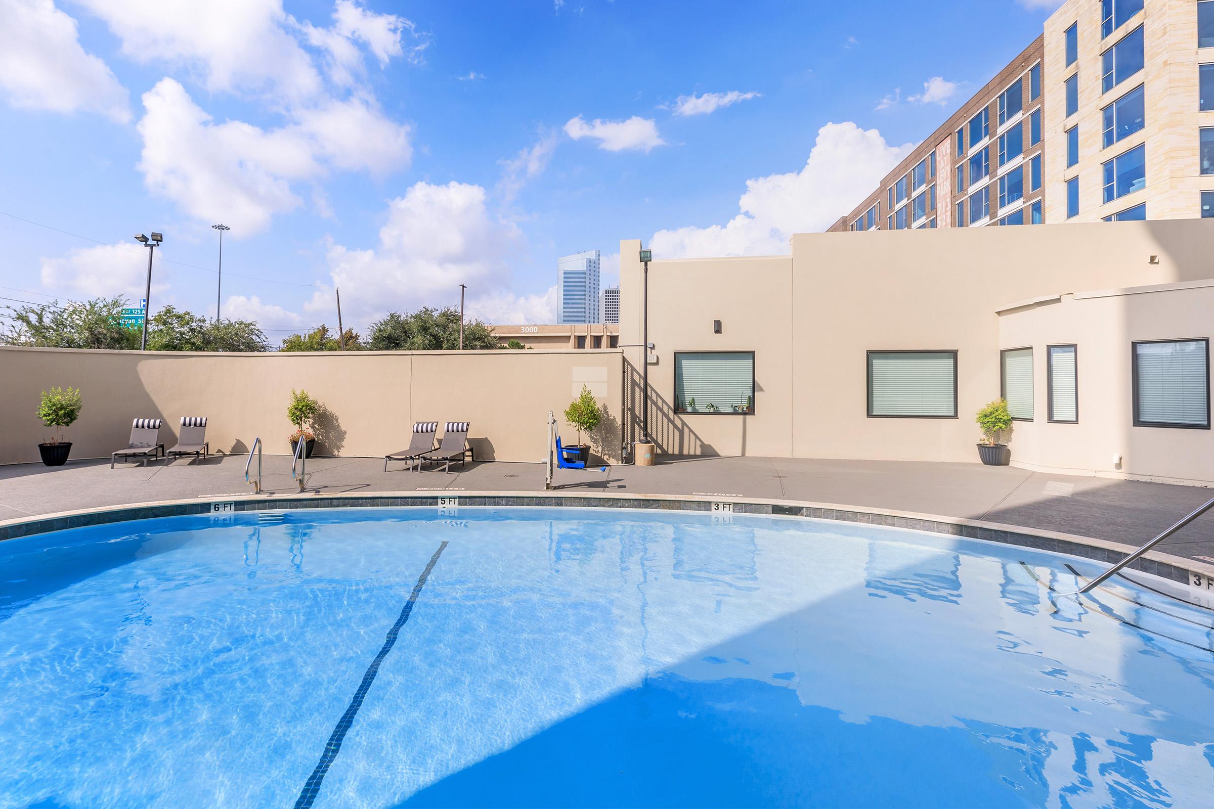 A clear blue swimming pool surrounded by lounge chairs, with a modern building in the background. The scene features a bright sky with fluffy clouds, creating a relaxing outdoor atmosphere.