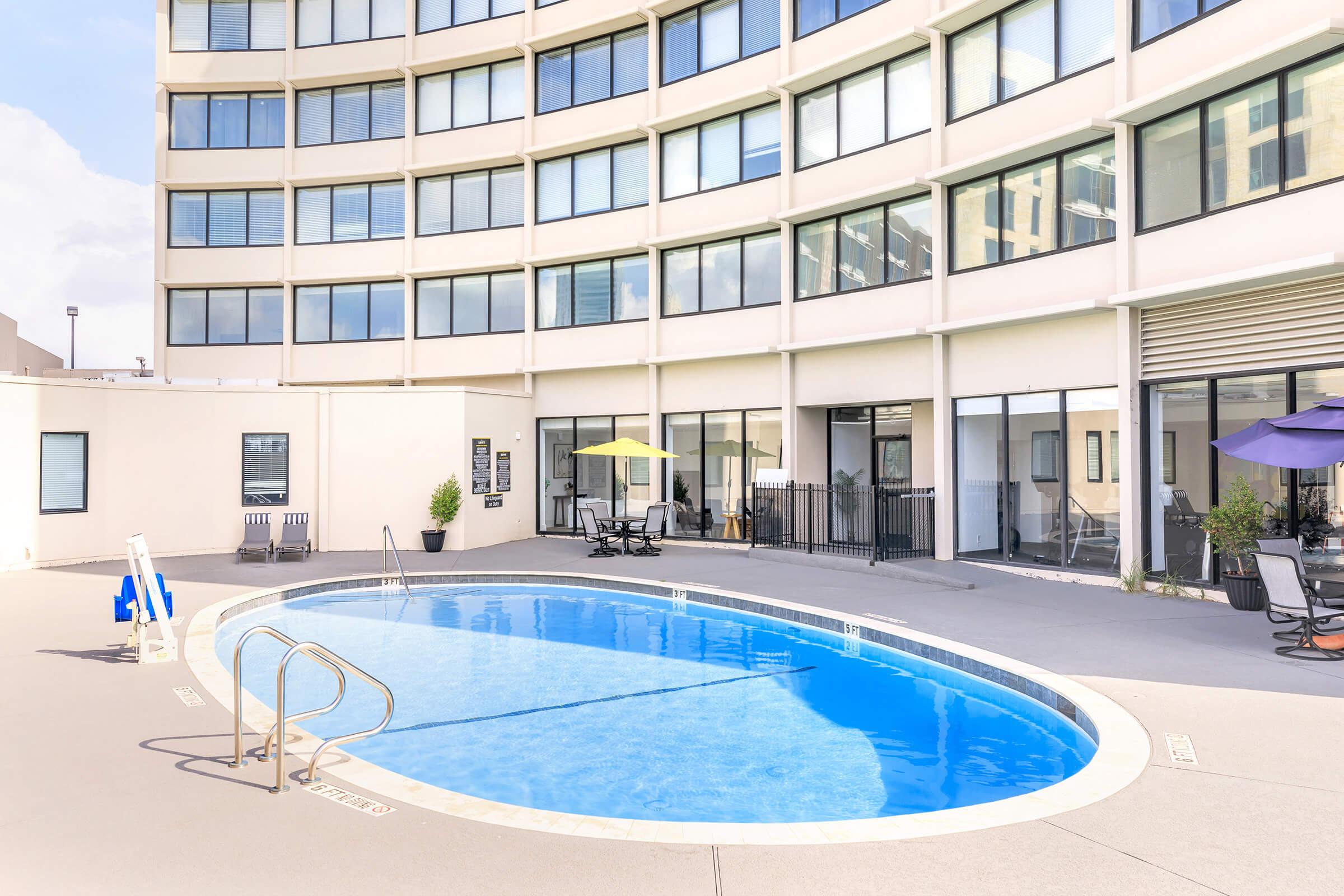 A modern outdoor pool area featuring a curved shape, surrounded by a large building with multiple windows. Lounge chairs and tables are positioned nearby, with umbrellas providing shade. The pool is clear blue, reflecting the sky, and there are potted plants adding greenery to the setting.