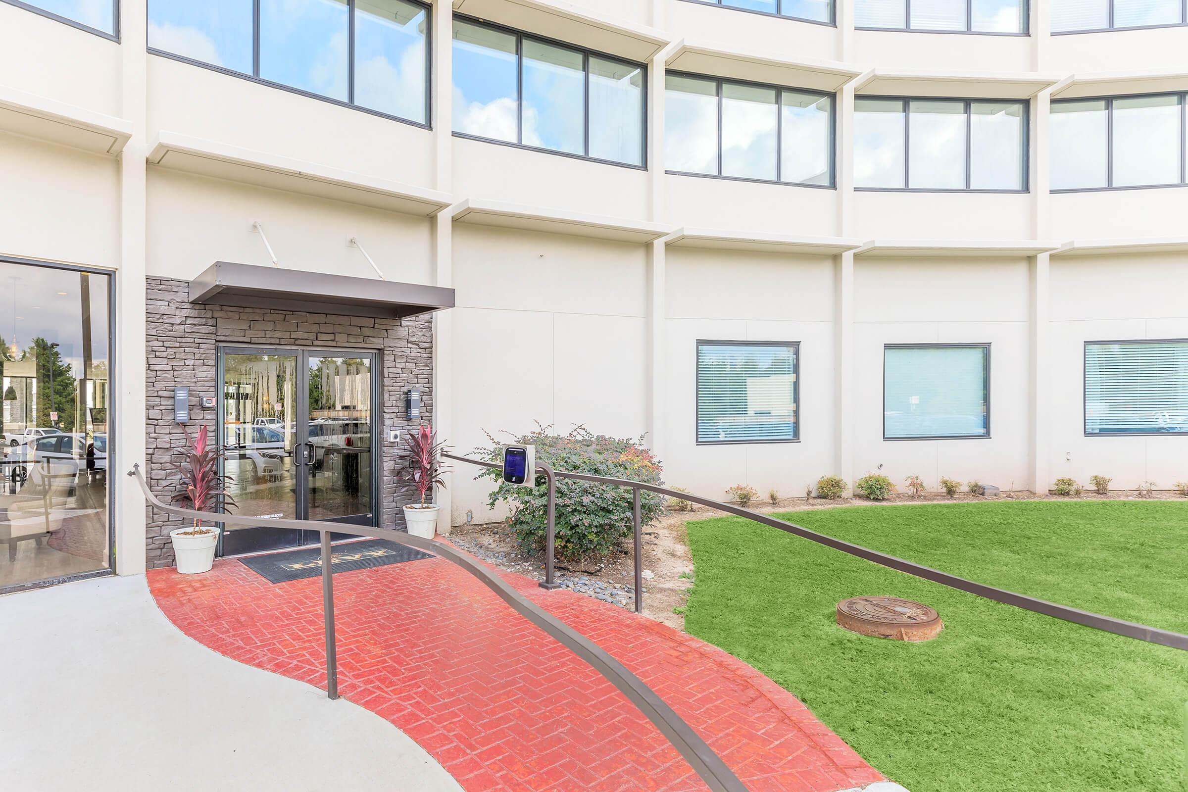 A modern building entrance featuring a curved, red brick pathway leading to double doors. The facade is light-colored with large windows, and there's green landscaping with potted plants in front. A small stone planter adds a natural touch to the entrance area.