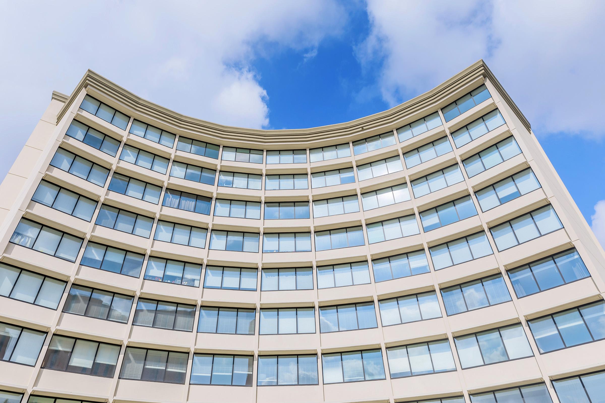 A modern building façade with a curved design, showcasing multiple rows of large windows against a backdrop of blue sky and scattered clouds. The structure features a light-colored exterior, emphasizing its sleek and contemporary architectural style.