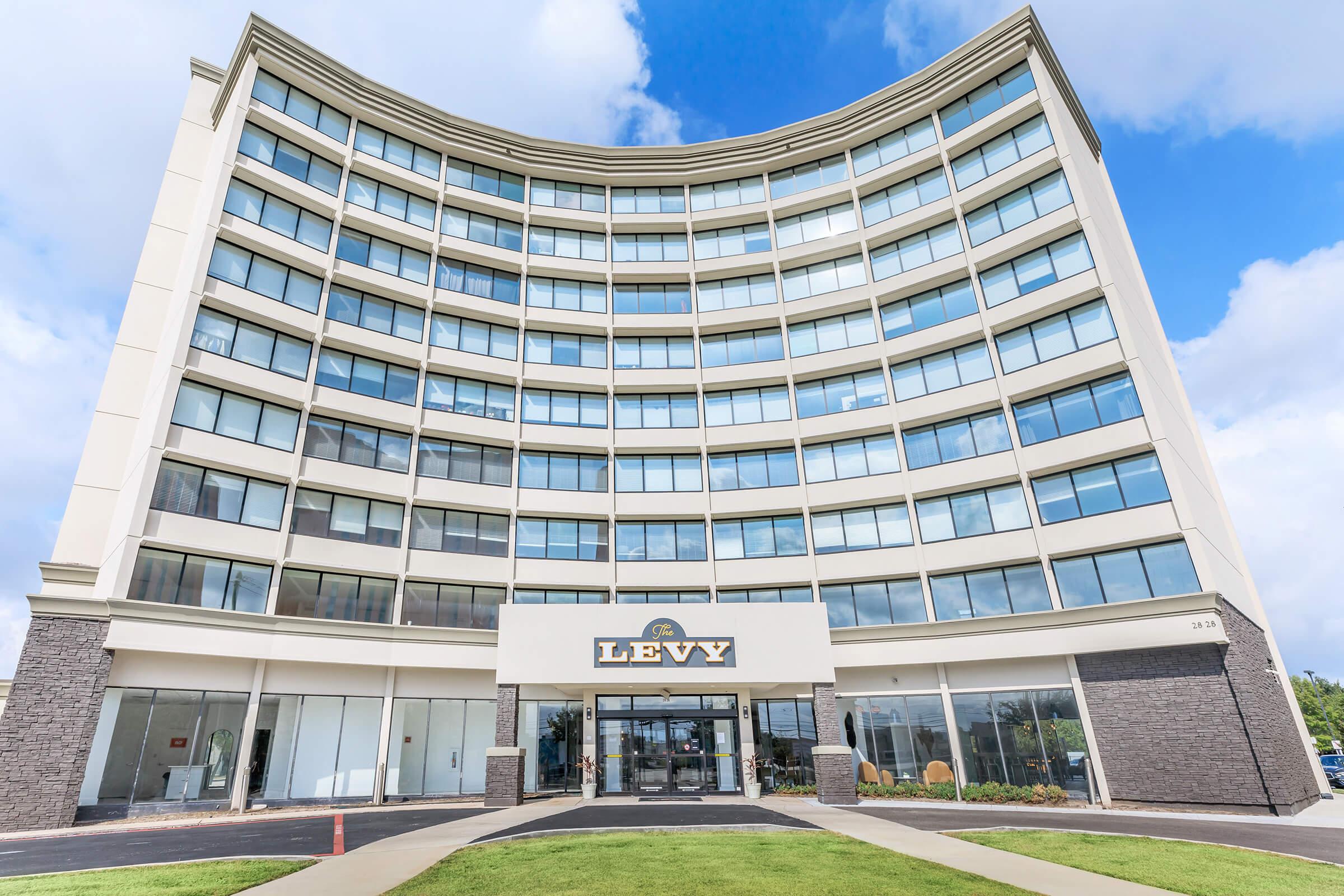 Modern multi-story building with a curved façade, featuring large windows. The entrance displays the name "LEVY" prominently. The surrounding area includes well-maintained grass and a paved walkway, under a partly cloudy sky.