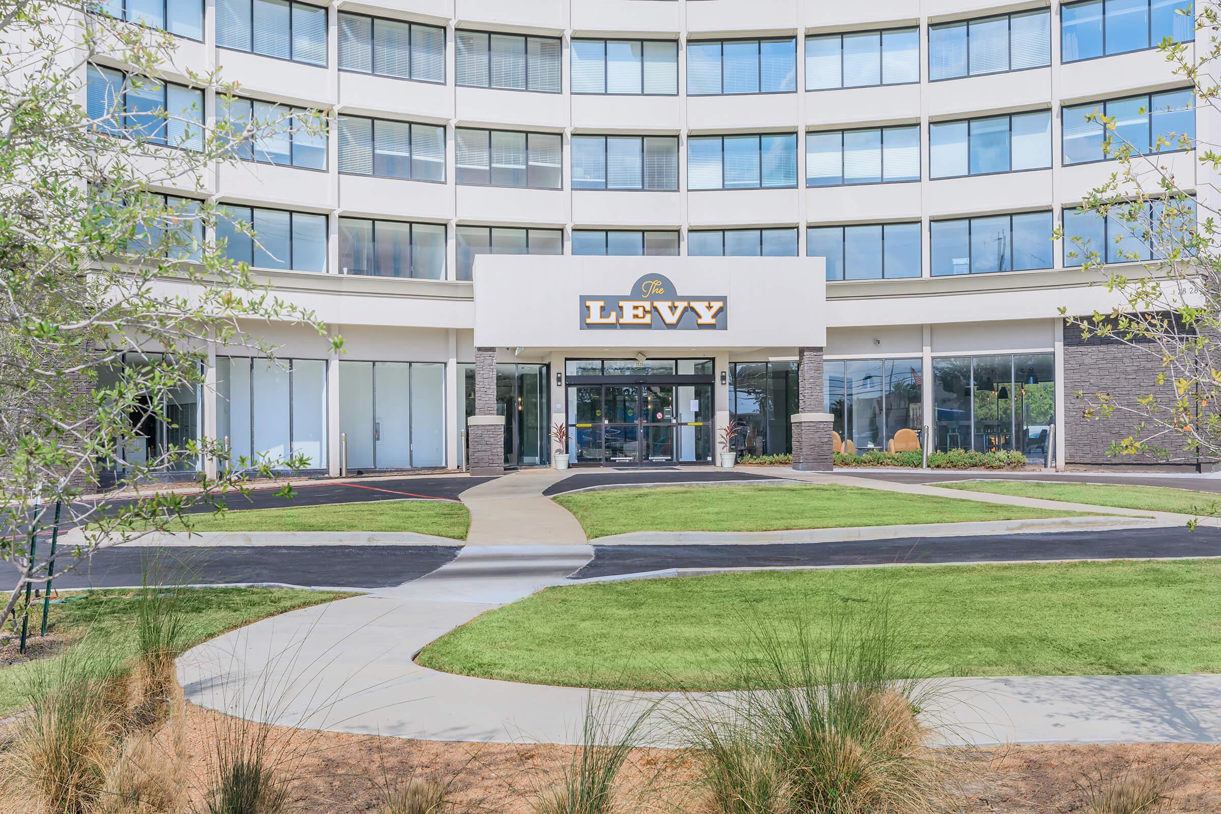 A modern building featuring a round architecture with large glass windows. The entrance is marked by a sign that reads "Levy." There are landscaped areas with green grass and pathways leading to the entrance, creating a welcoming atmosphere.